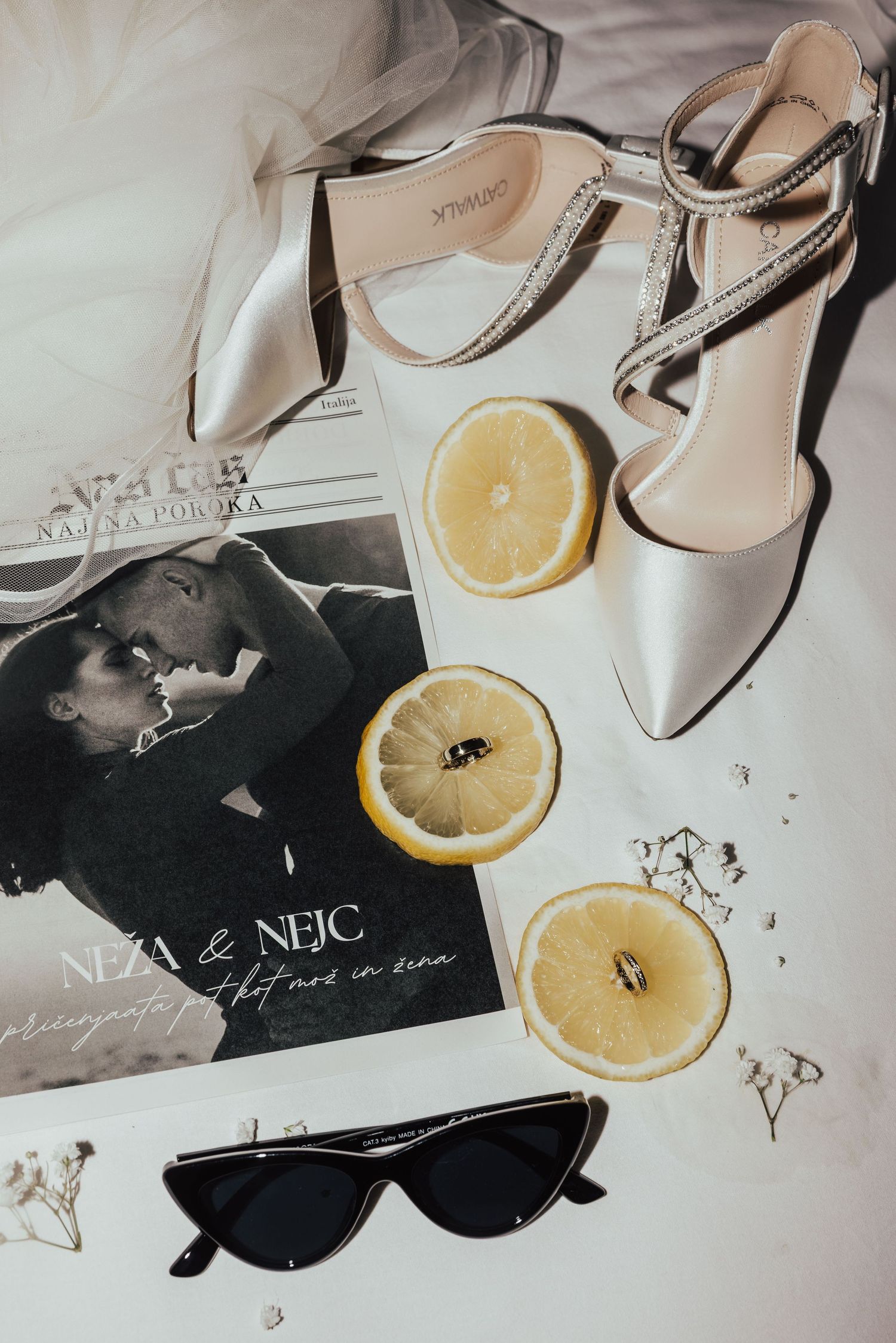 Elegant wedding flat lay with white heels, lemon slices, rings, sunglasses, and a couple's magazine on white fabric.