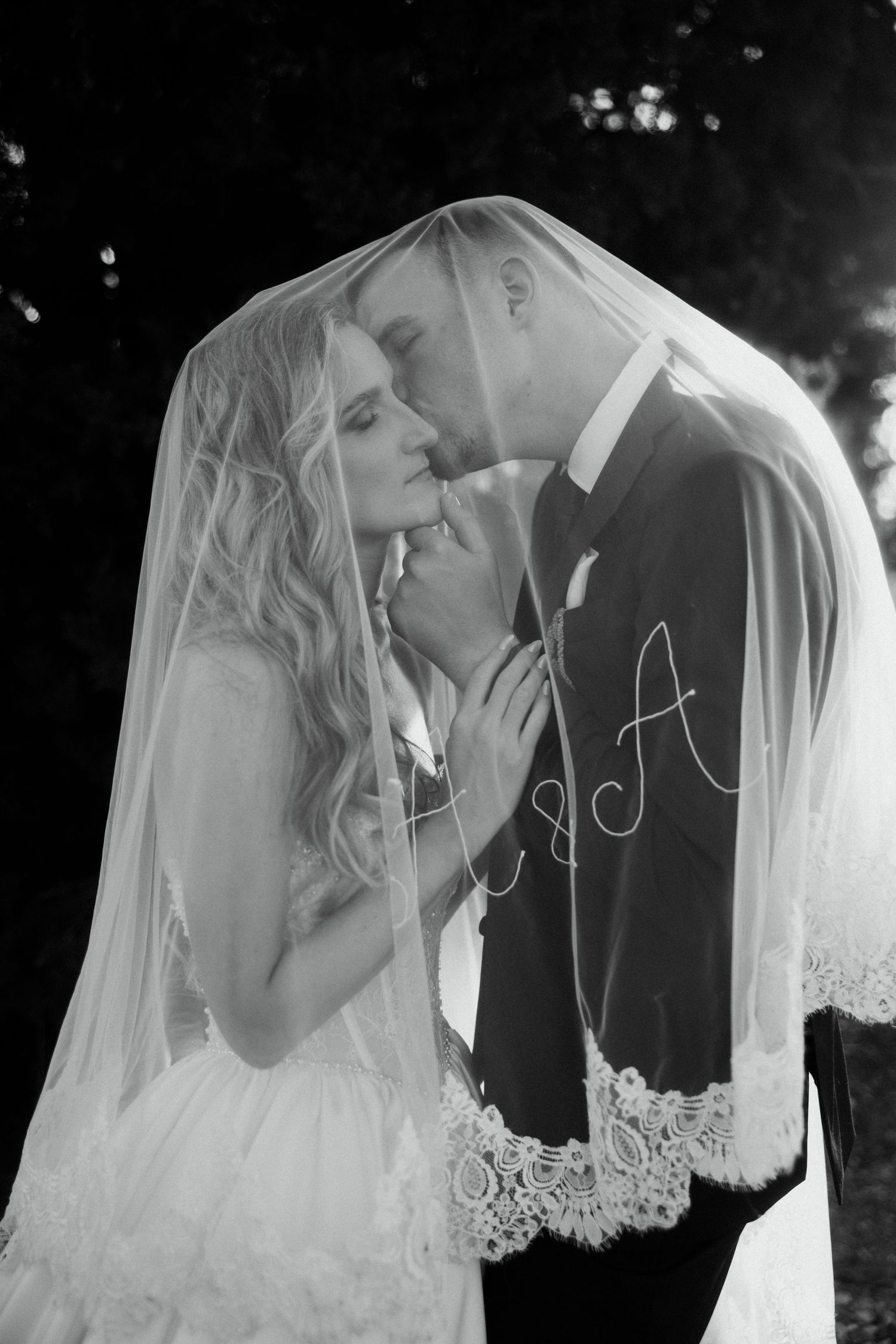 Black and white photo of bride and groom sharing a romantic moment under a lace veil with monogram.