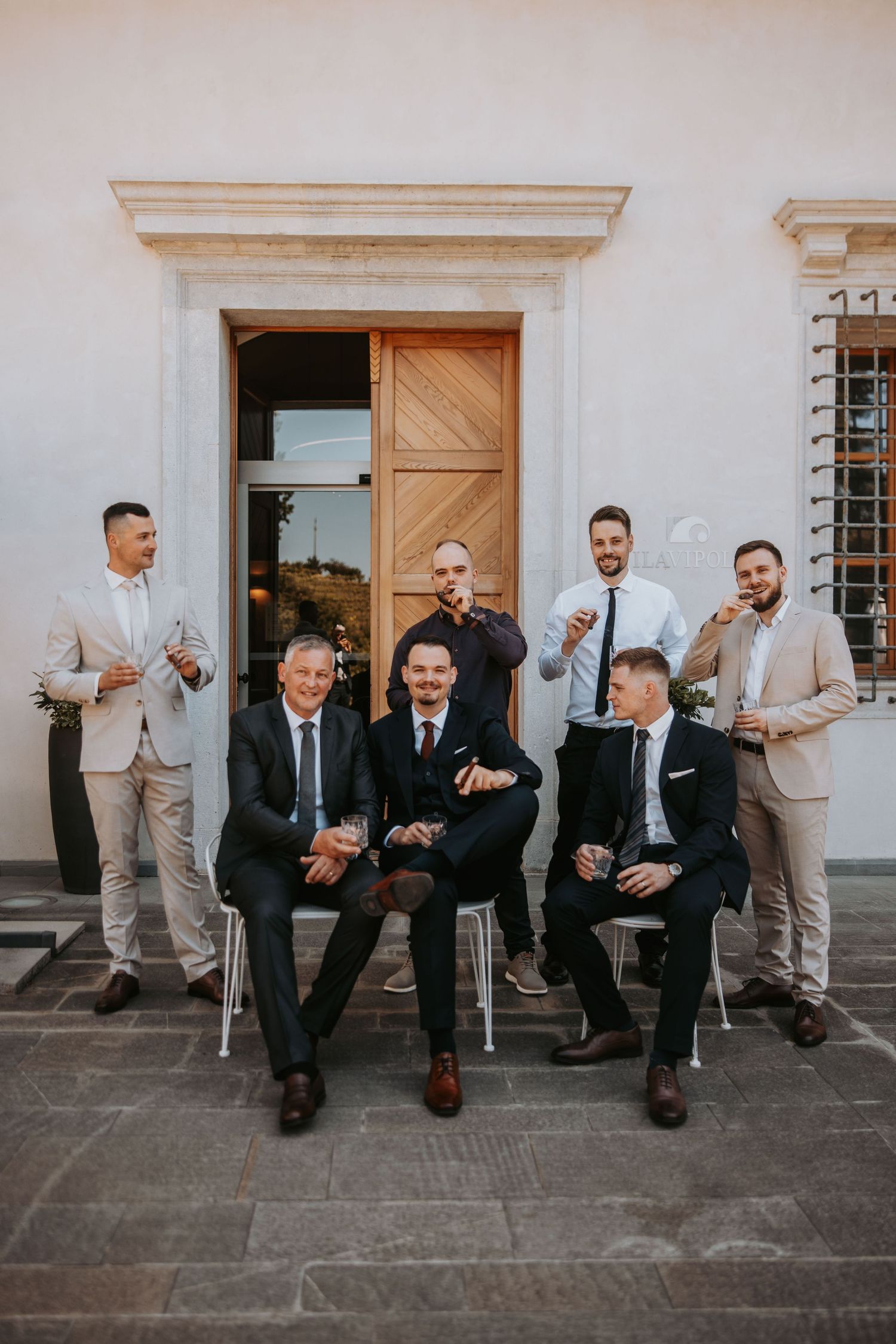 Groom and groomsmen posing in suits outside a European villa, some seated and holding drinks.
