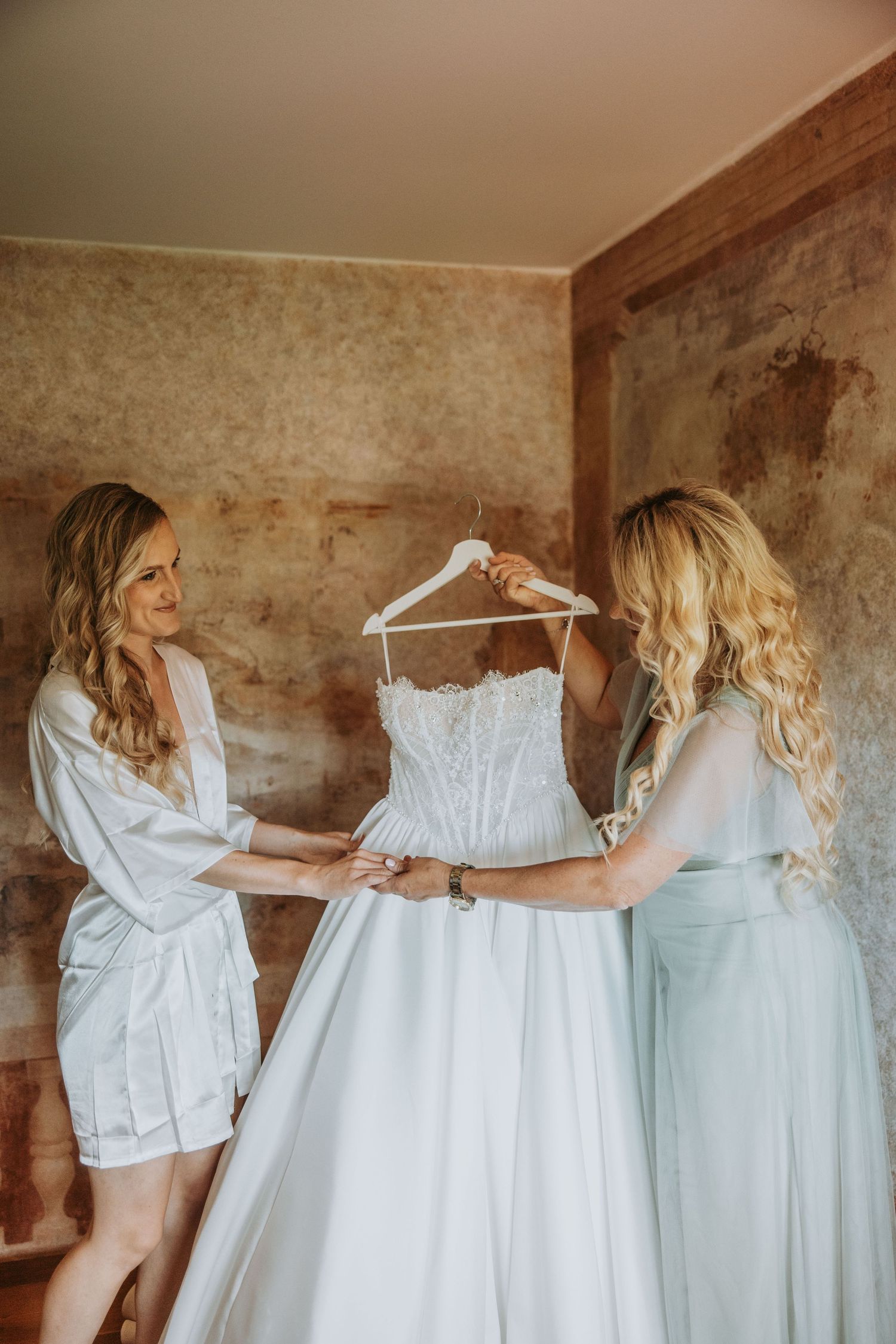 Bride in white robe holding wedding gown on hanger with bridesmaid in rustic room.