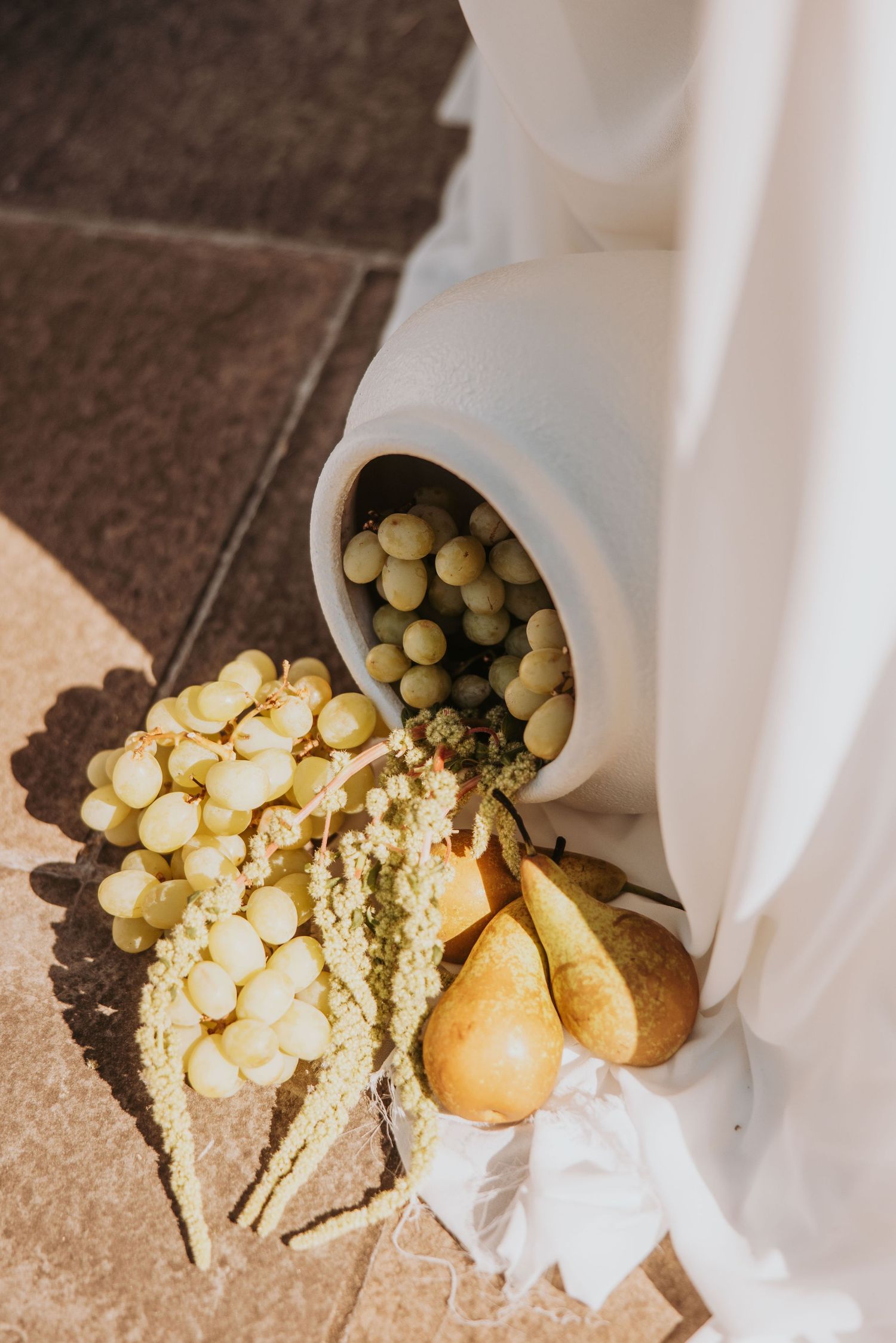 Elegant wedding decor featuring white ceramic vase with green grapes, pears, and dried wheat on tiled floor.