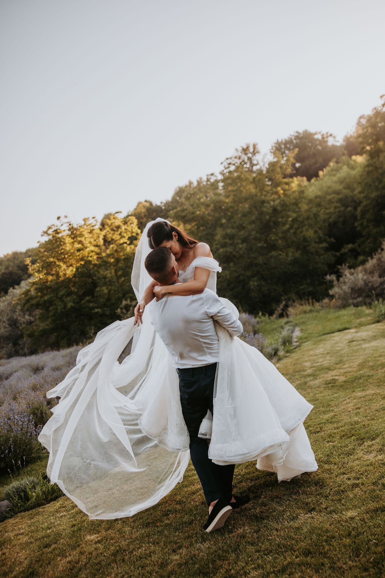 Groom lifts and spins bride in white wedding dress outdoors during golden hour celebration.