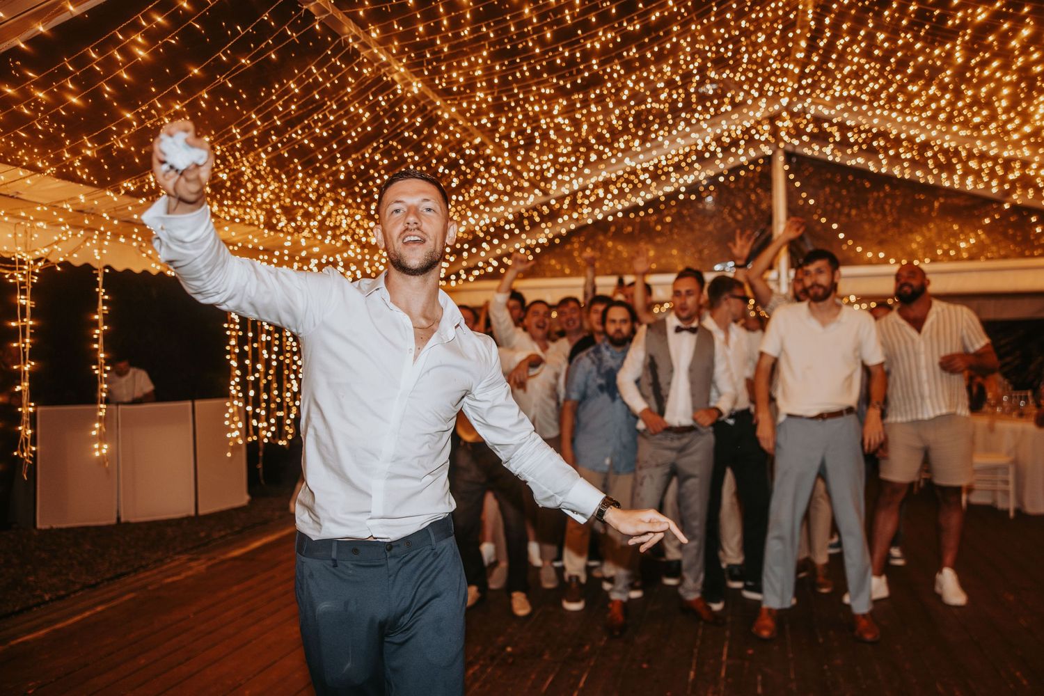 Groom tossing garter on dance floor under fairy lights surrounded by cheering wedding guests.
