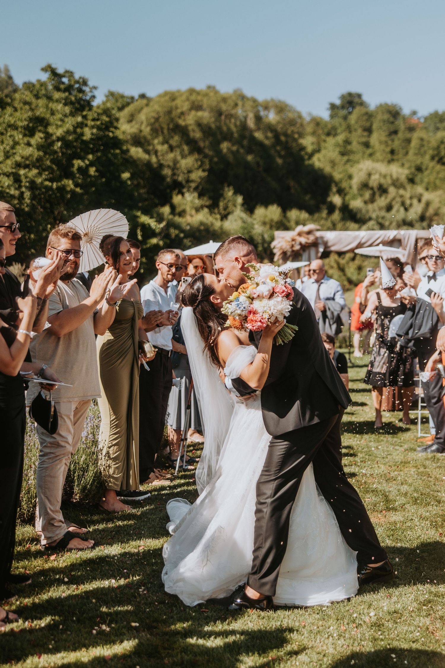 Newlyweds share a romantic kiss outdoors as wedding guests celebrate around them on a sunny day.