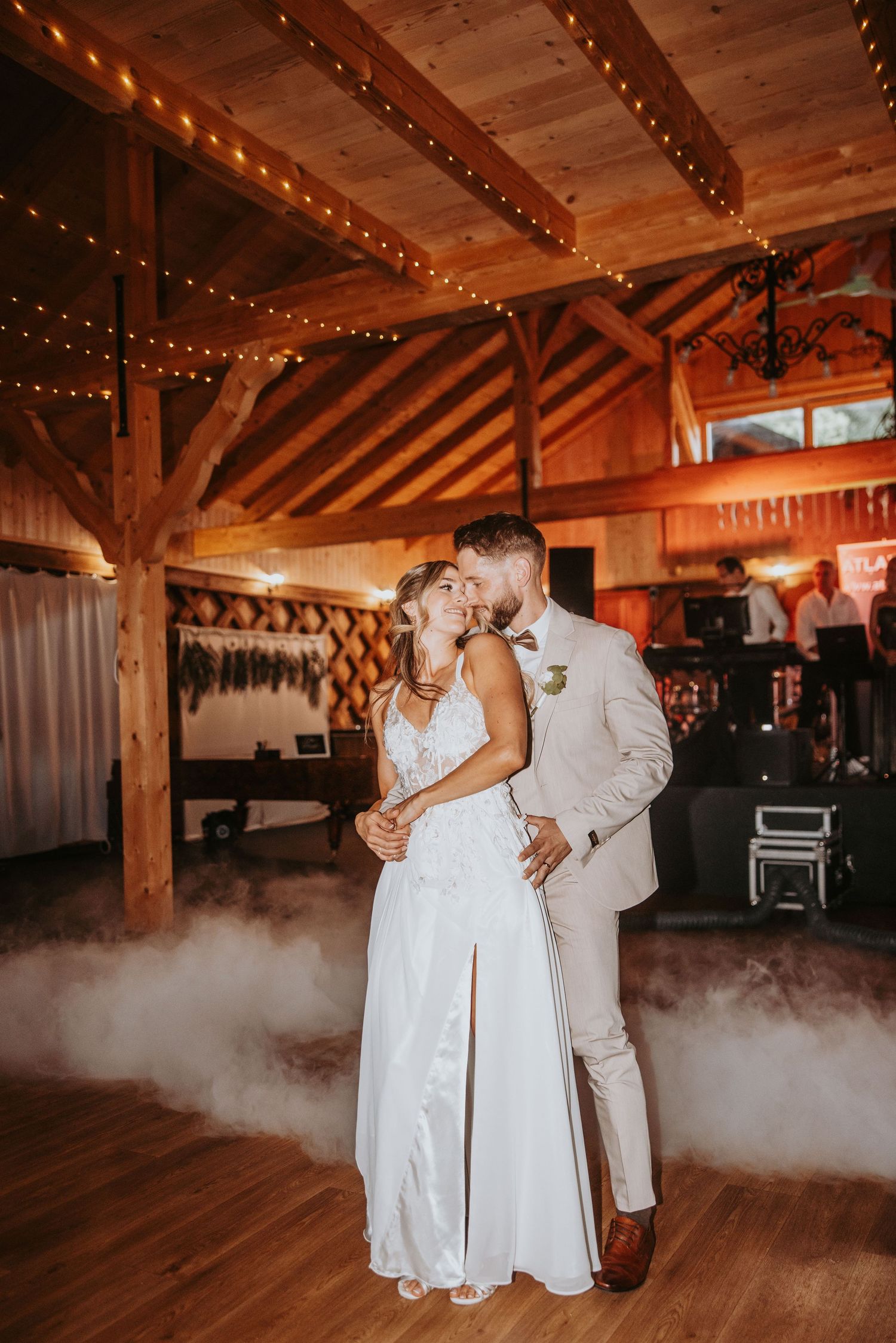 Bride and groom share first dance in rustic barn venue with string lights and romantic fog effect.