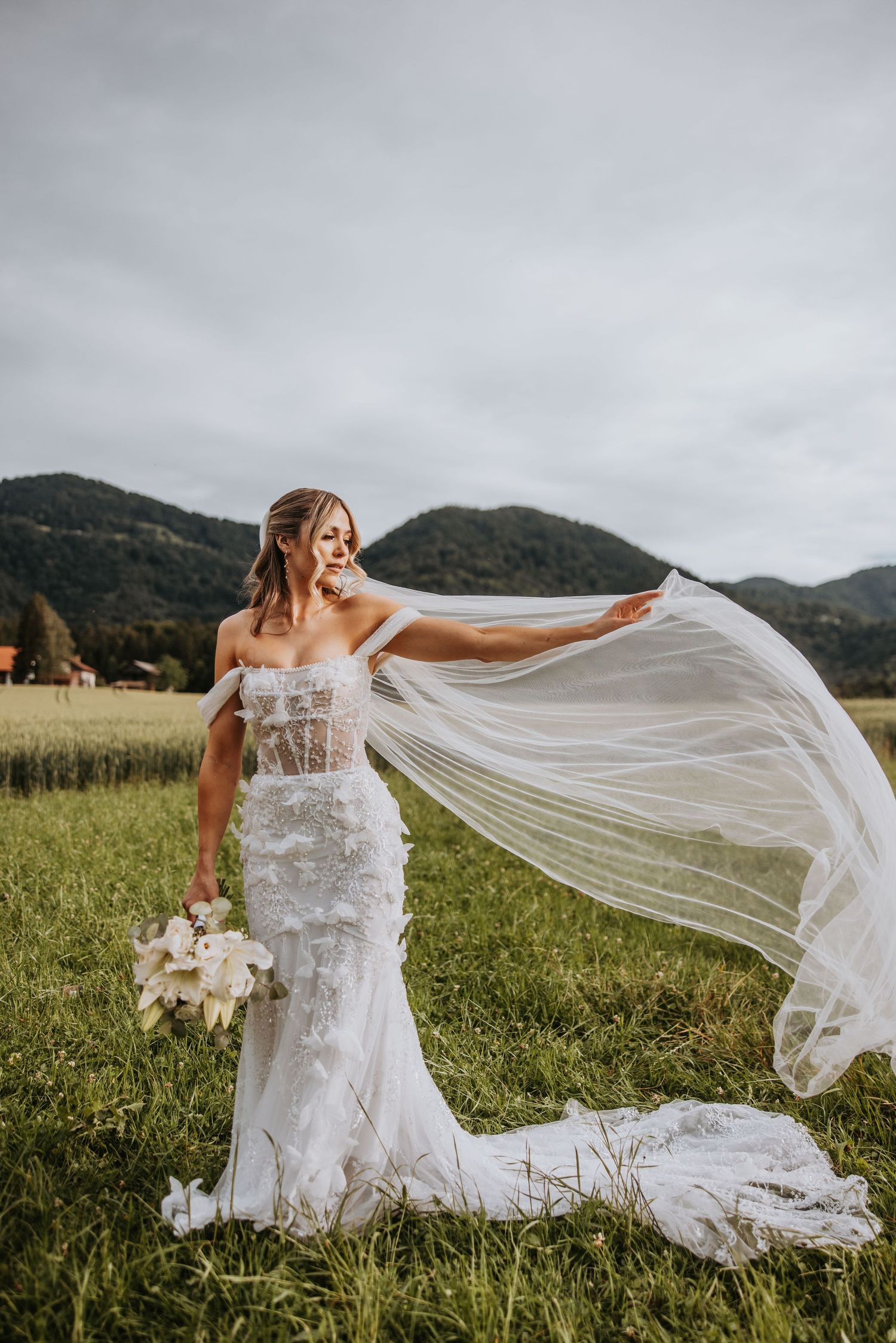 Bride in lace mermaid gown extends flowing veil in mountain meadow holding white floral bouquet.