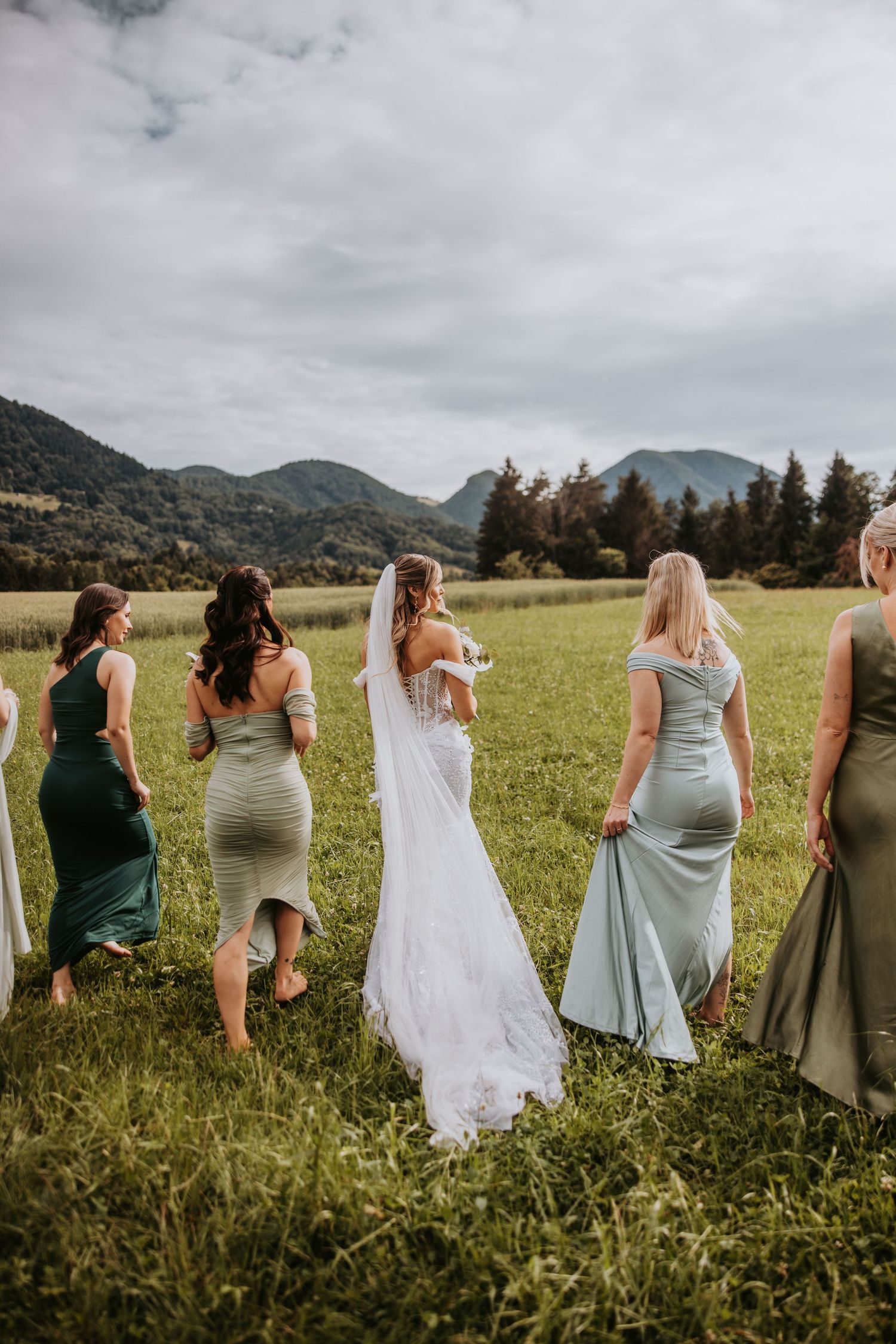 Bride and bridesmaids in green dresses walking through a meadow with mountain views on a wedding day.