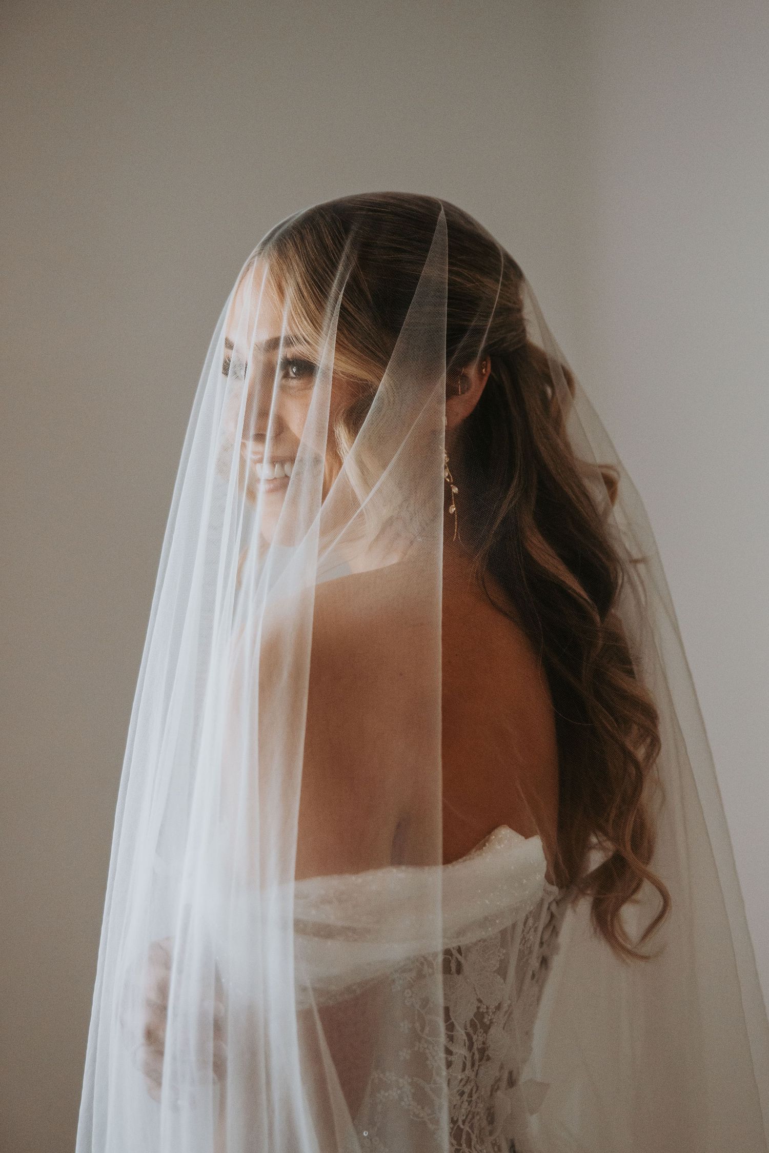 Bride with curly hair smiling softly while draped in a flowing white wedding veil from behind.