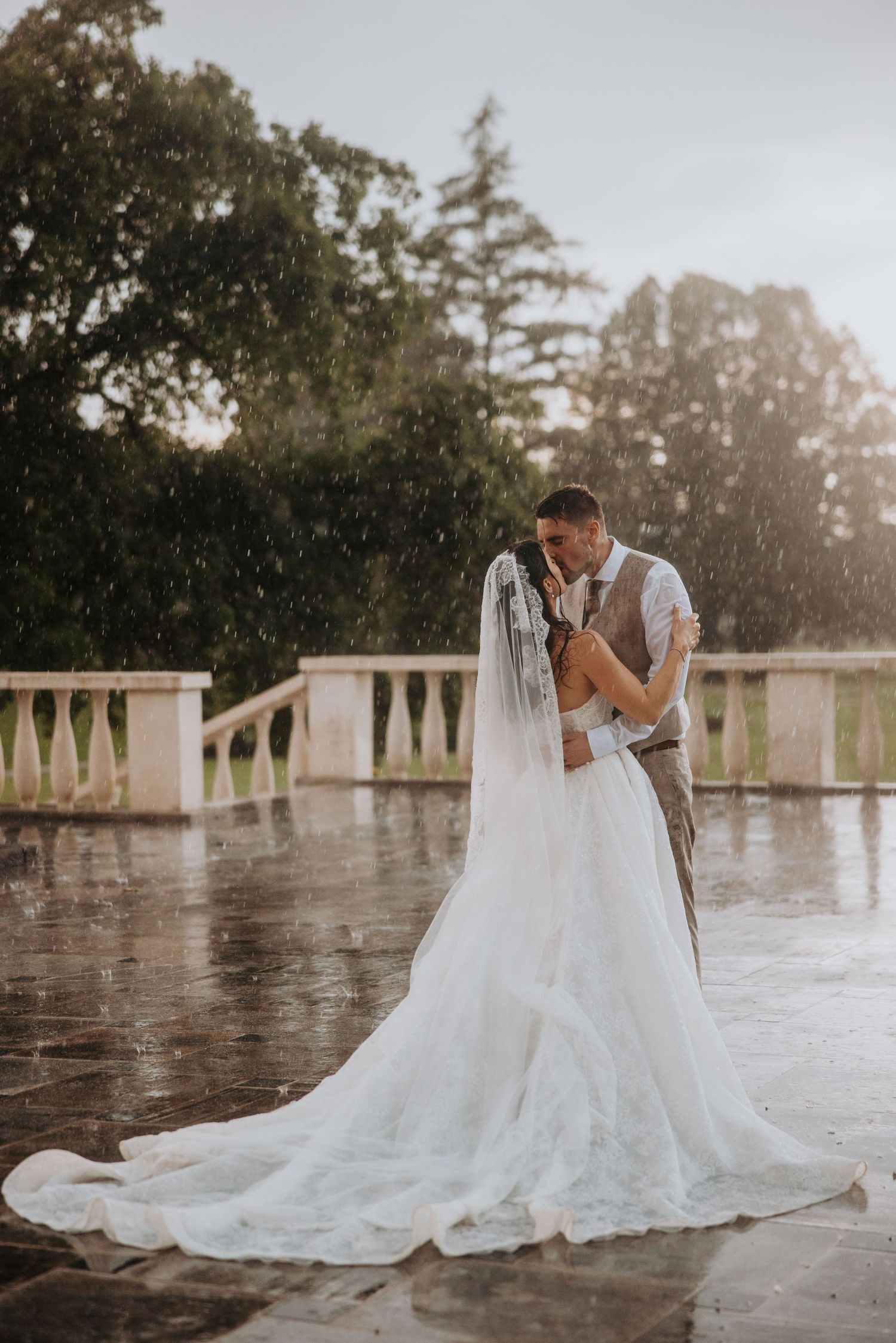Romantic couple sharing a kiss in the rain, bride in a lace gown with long train on a stone terrace.