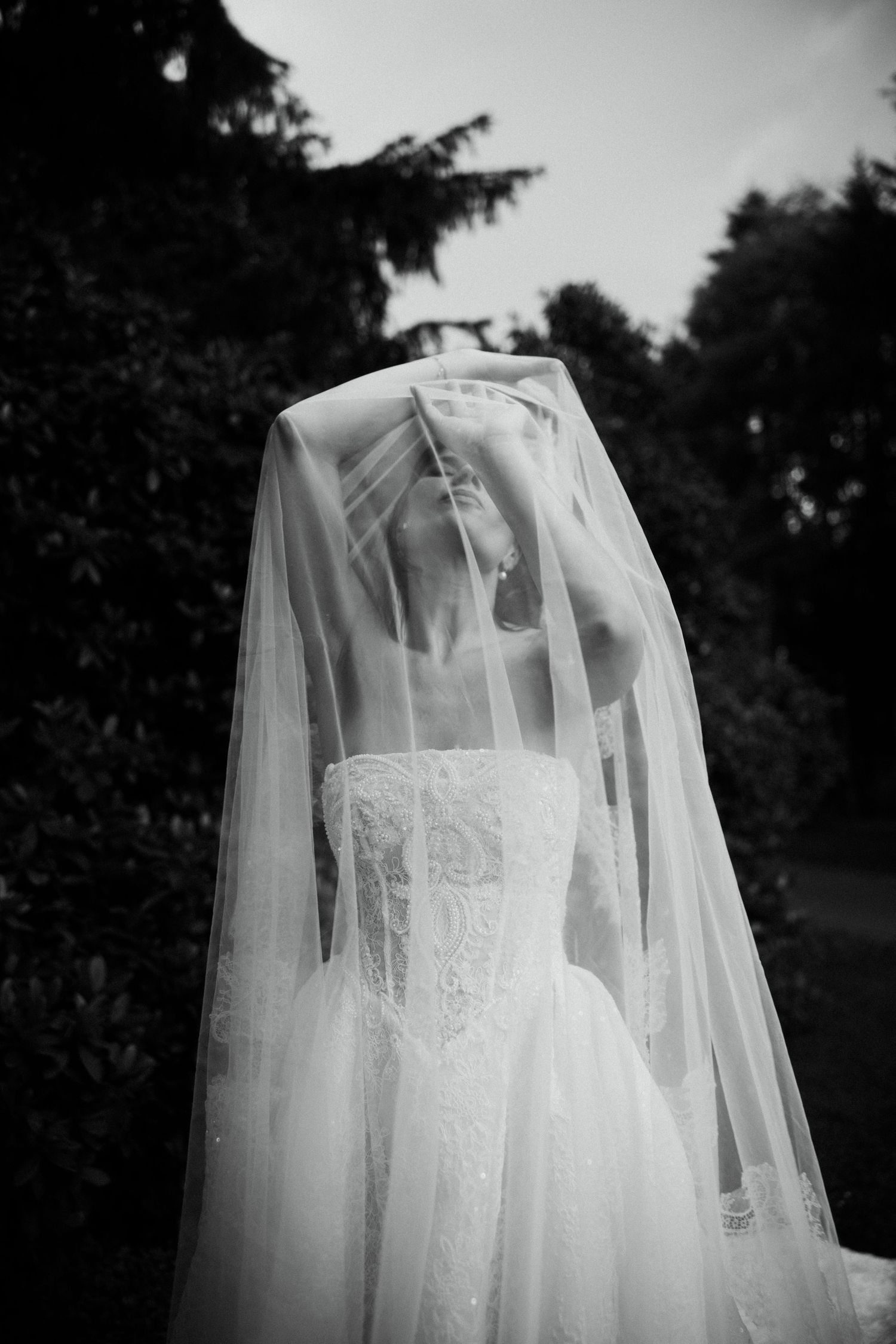 Elegant bride in lace wedding dress lifts flowing veil overhead in dreamy black and white portrait.
