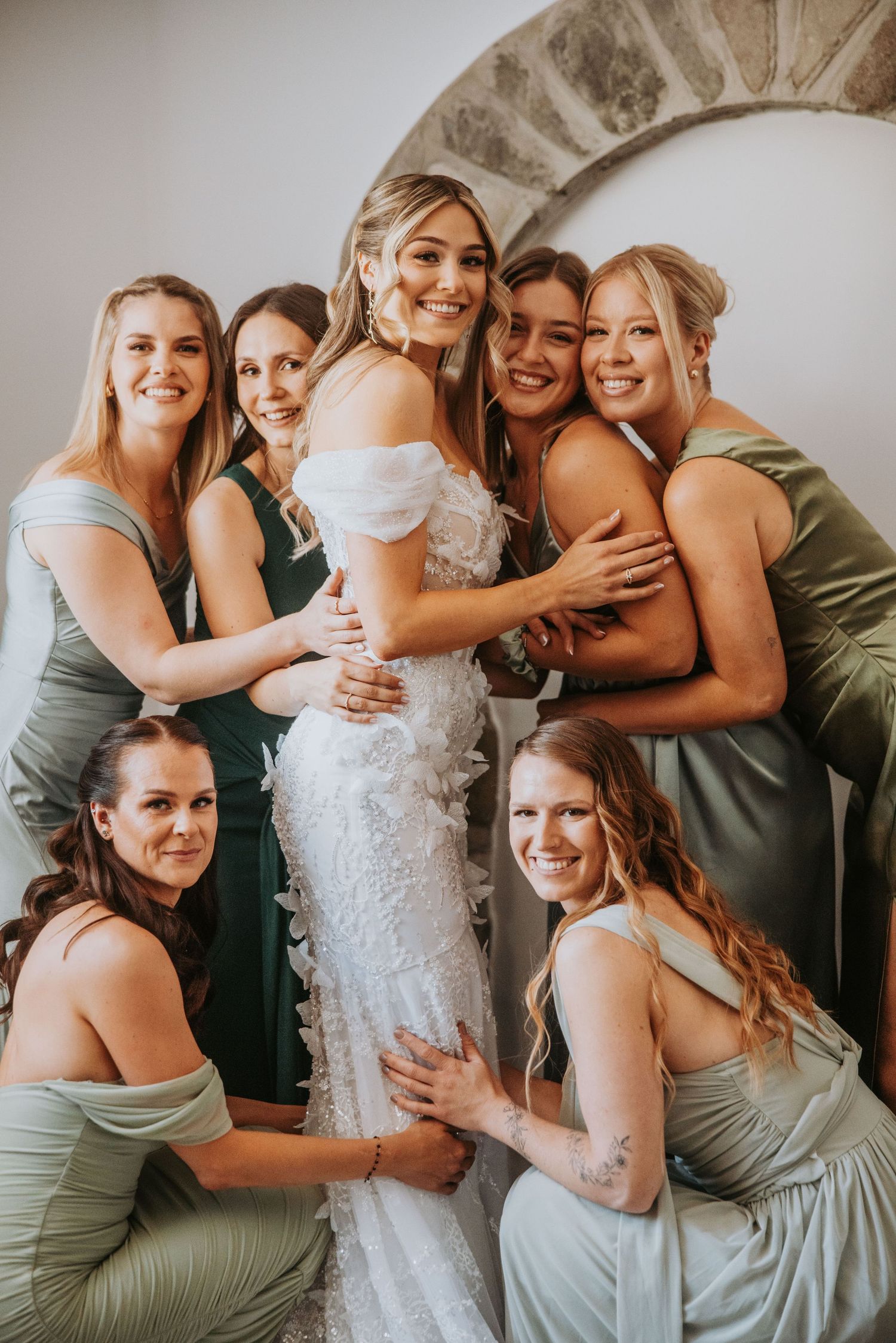 Smiling bride in lace wedding dress posing with six bridesmaids in sage and green gowns.