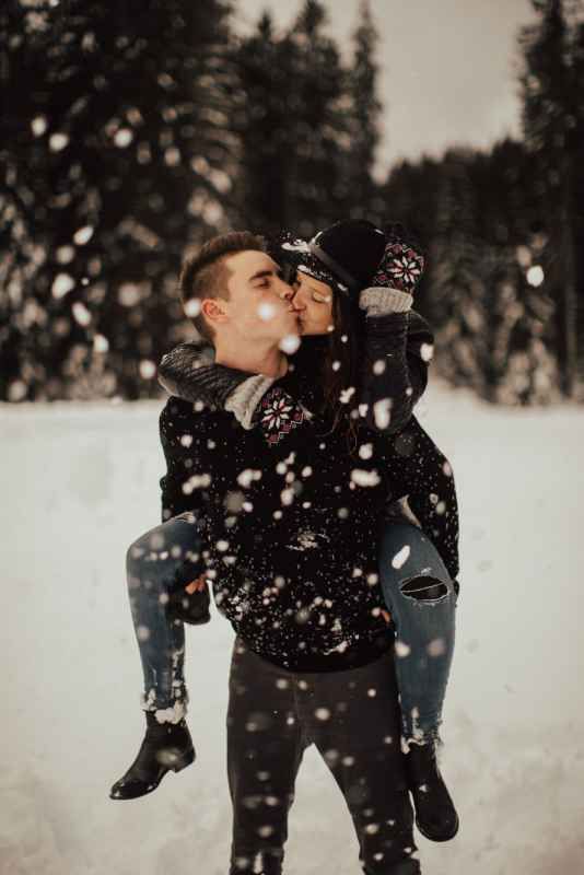 Couple sharing a kiss in snowy winter forest, man giving woman a piggyback ride.