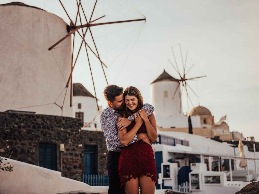 Couple embracing romantically in Santorini Greece with iconic white windmills and Cycladic architecture in background.