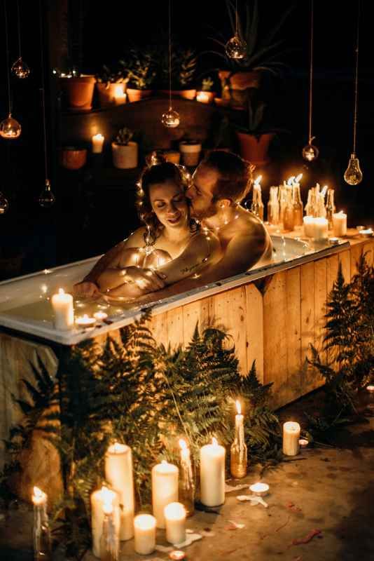 Romantic couple relaxing in a candlelit bath surrounded by ferns and glowing string lights in a cozy setting.