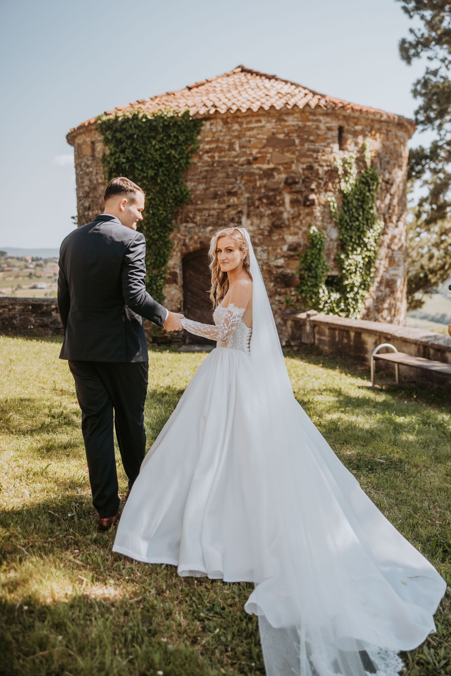 Bride in elegant ballgown with lace bodice and long veil holds grooms hand near ivy-covered stone chapel outdoors.