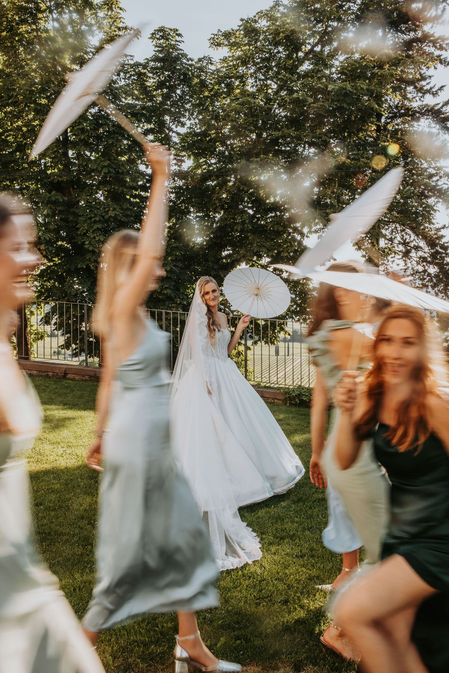 Bride in white gown dances joyfully with bridesmaids holding parasols on green lawn in golden sunlight.