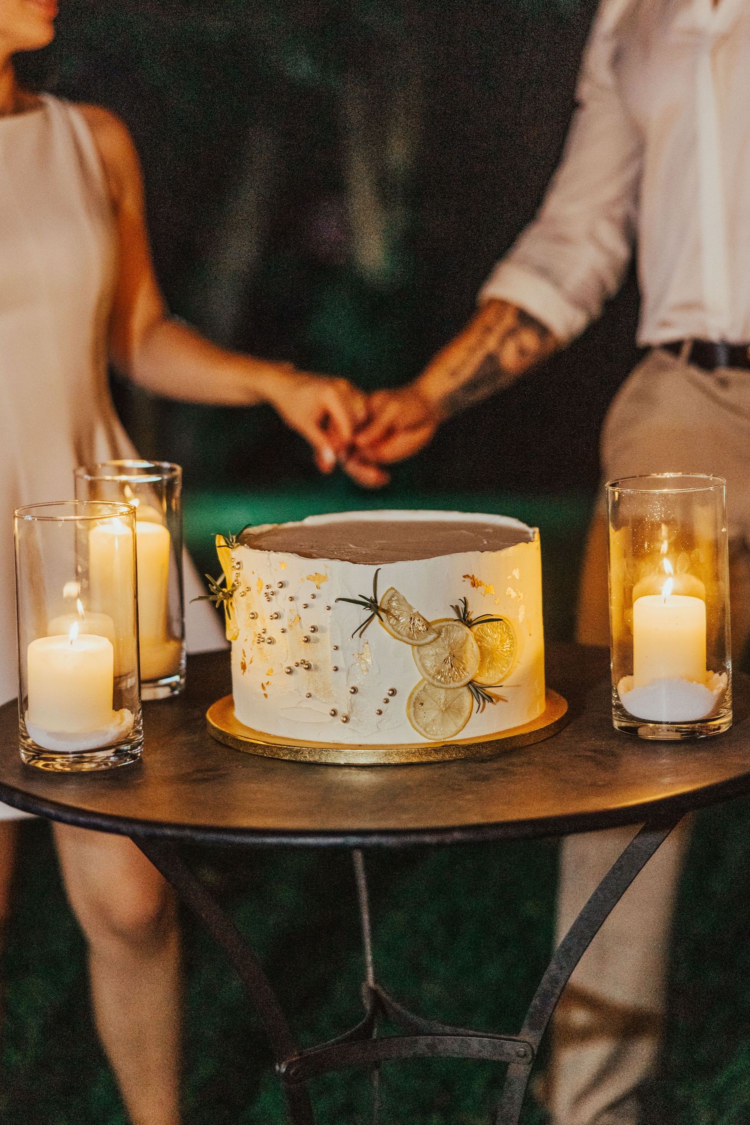 Elegant wedding cake with lemon botanical design surrounded by glowing candles on a gold table with couple holding hands.