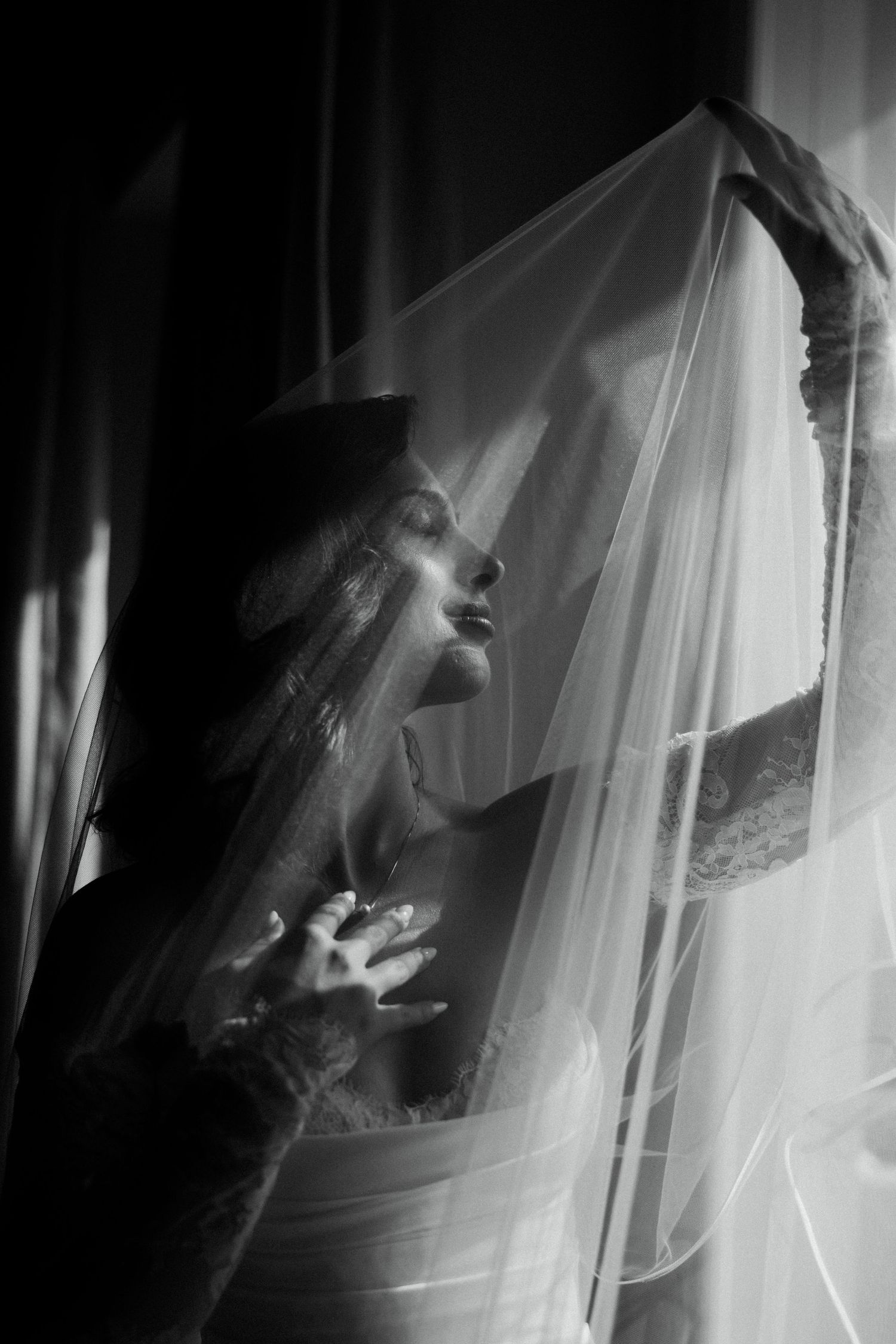 Dramatic black and white portrait of a bride lifting her veil in soft window light.