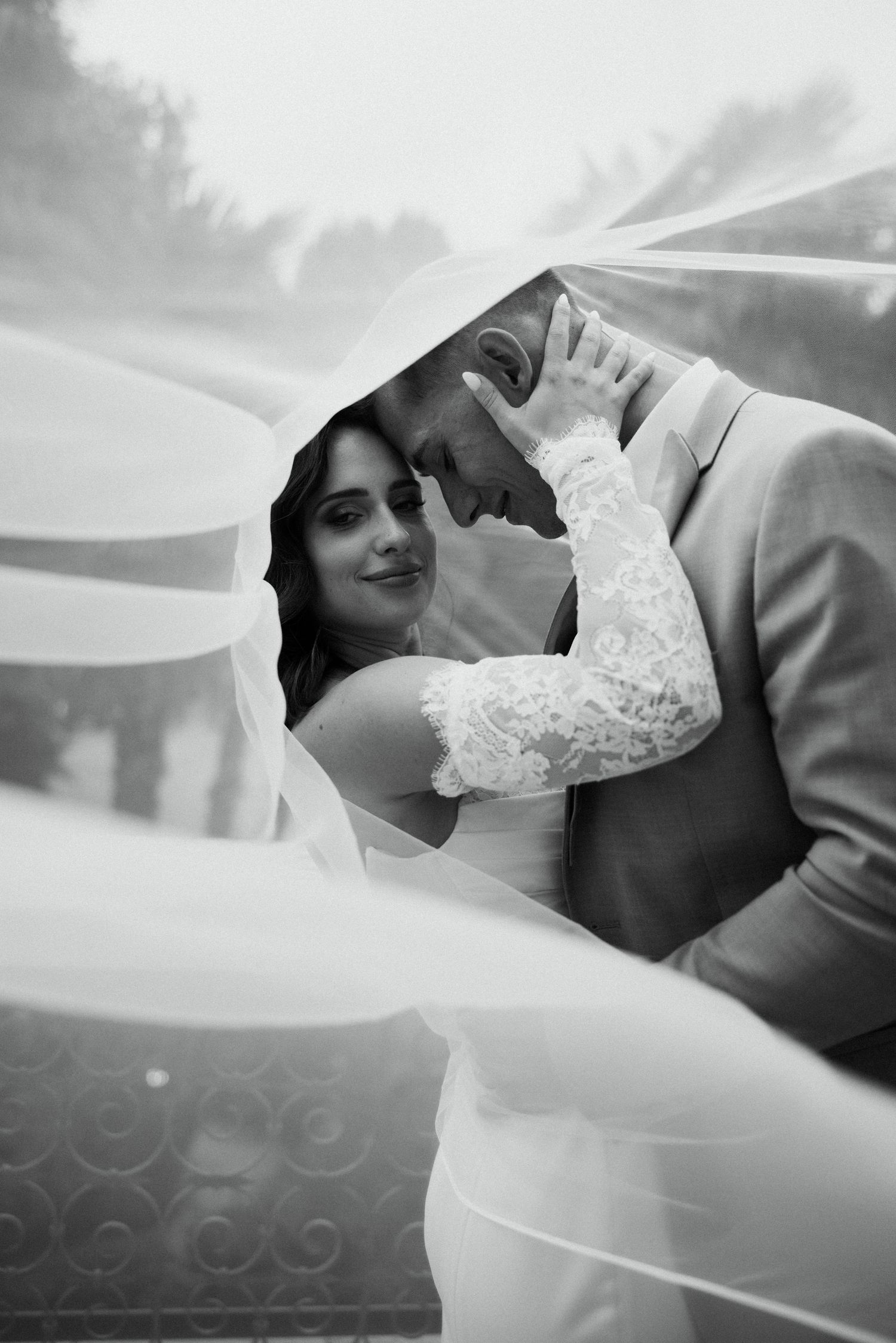 Romantic black and white photo of couple embracing under flowing bridal veil.
