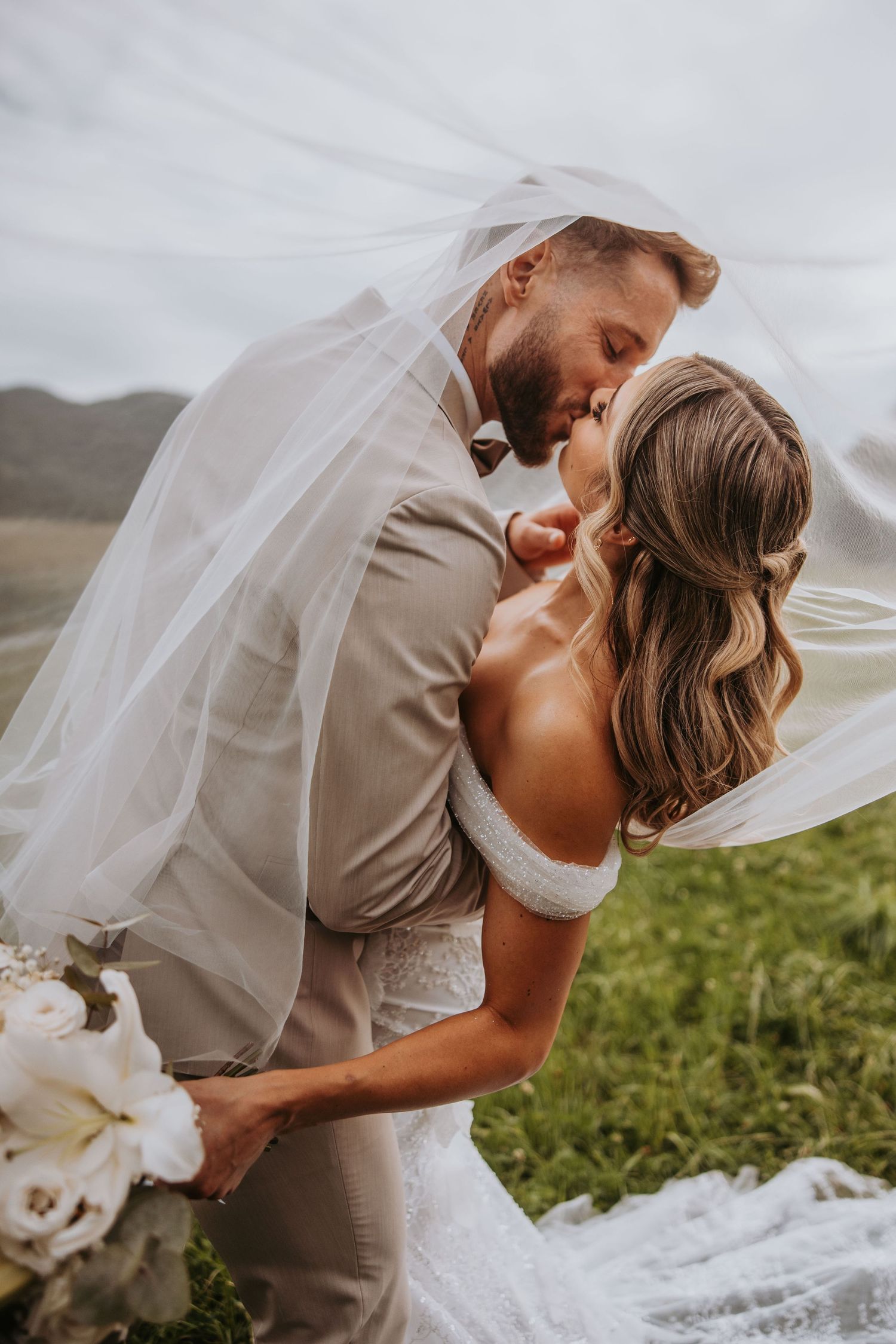 Groom dipping bride under flowing veil for romantic kiss in lush green outdoor setting.
