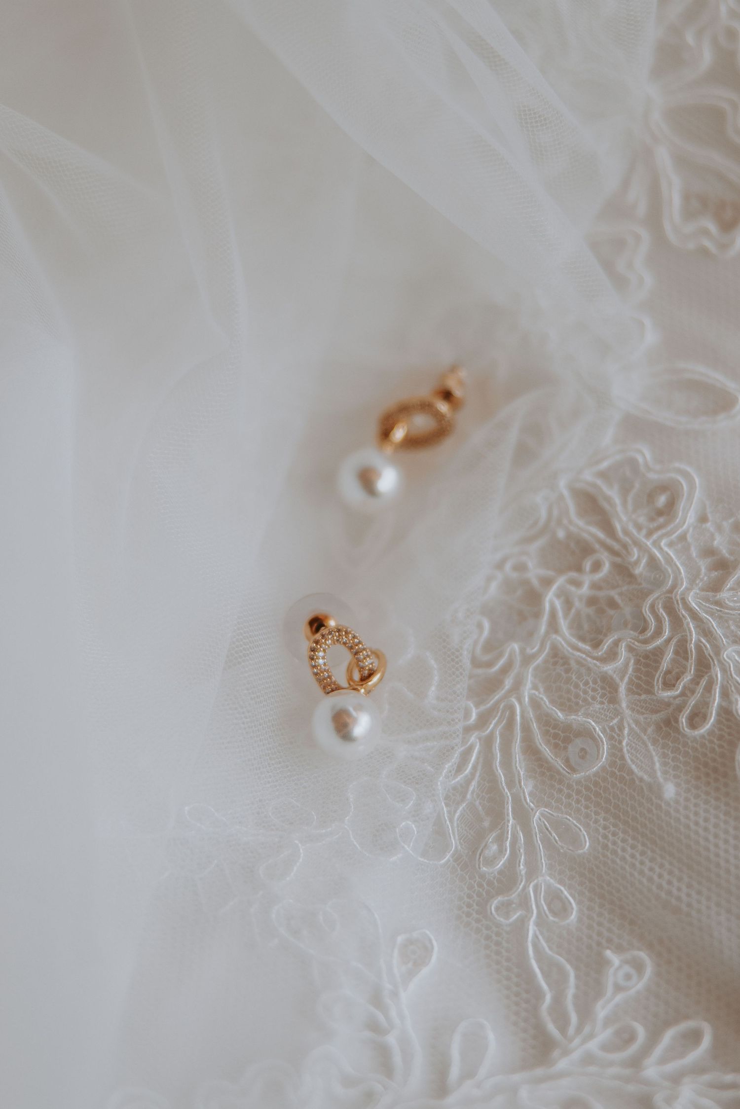 Gold and pearl drop earrings with diamond accents resting on white lace bridal veil fabric.