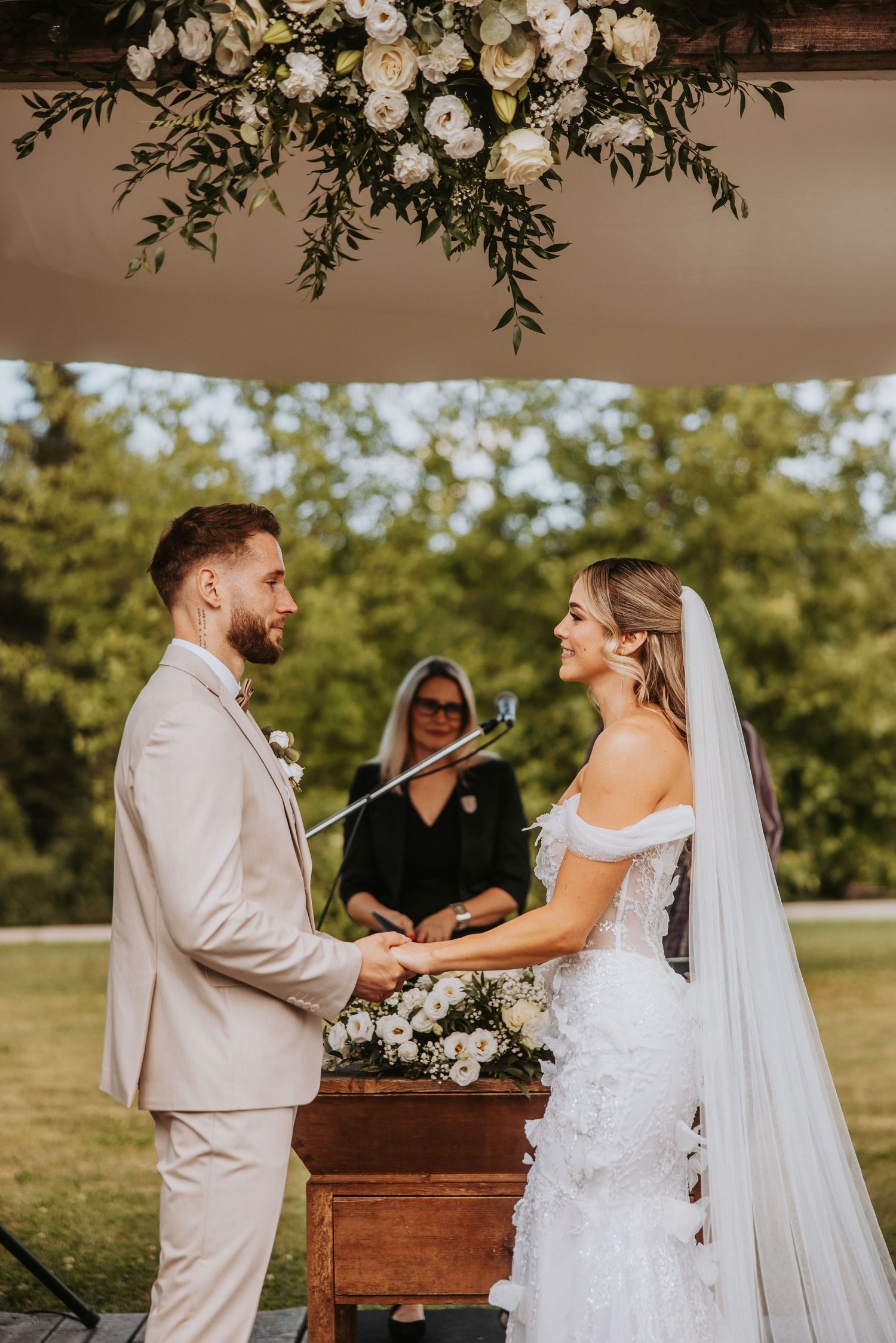 Bride and groom holding hands at floral-decorated outdoor wedding ceremony with officiant in background.