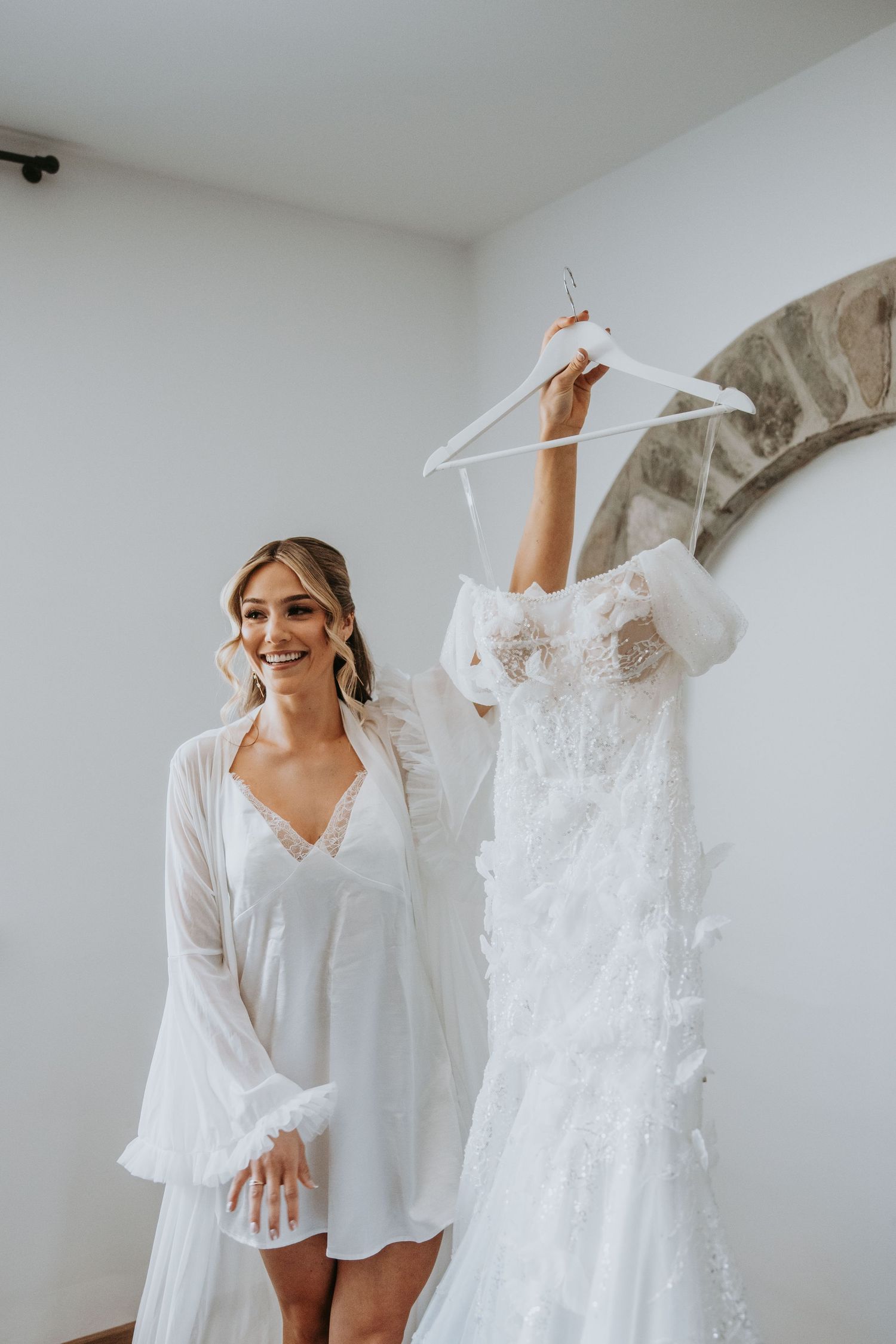 Smiling bride in white robe holds up elegant lace wedding dress on hanger in bright minimalist room.
