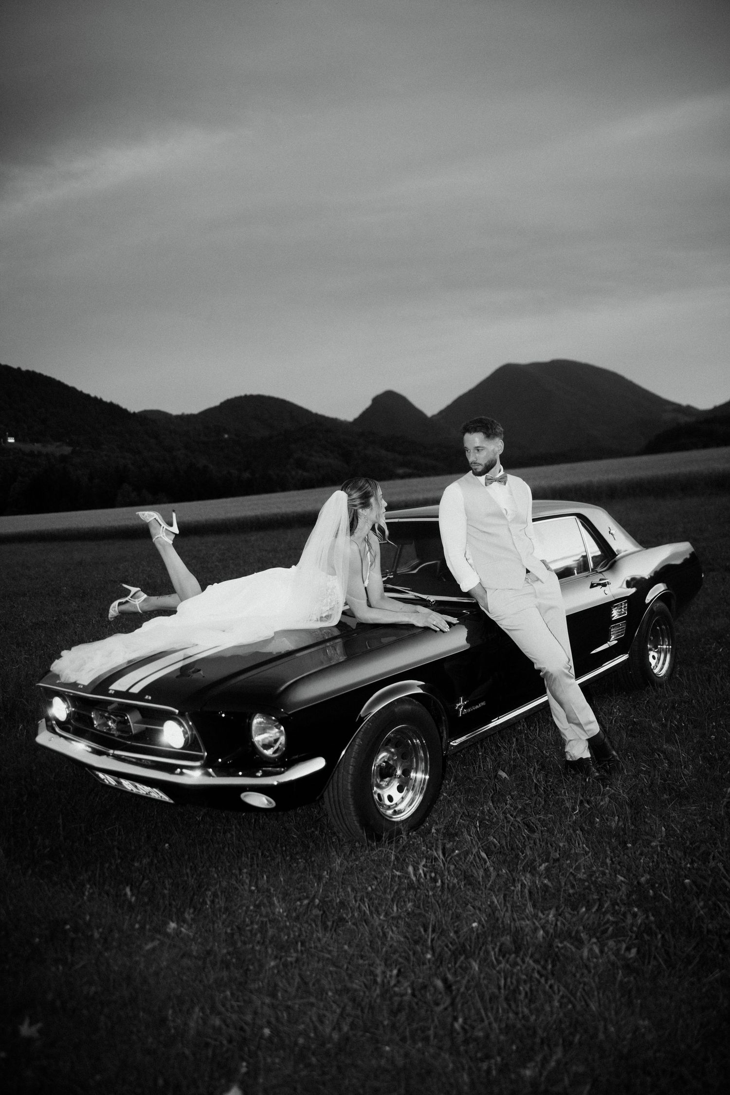 Bride and groom pose with a vintage Ford Mustang in a scenic mountain field in black and white.