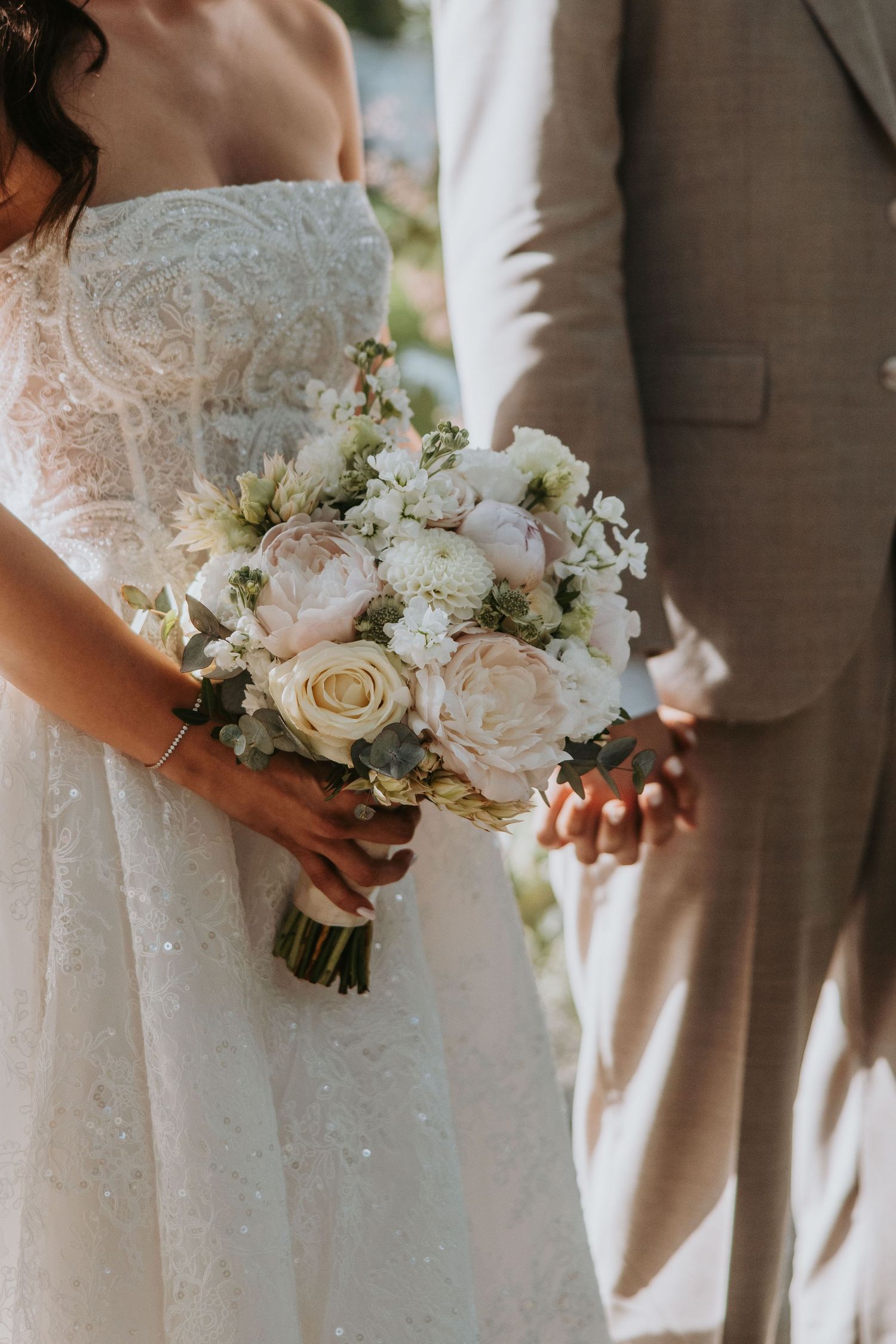 Bride in lace gown holds romantic blush and white peony bouquet while holding grooms hand.