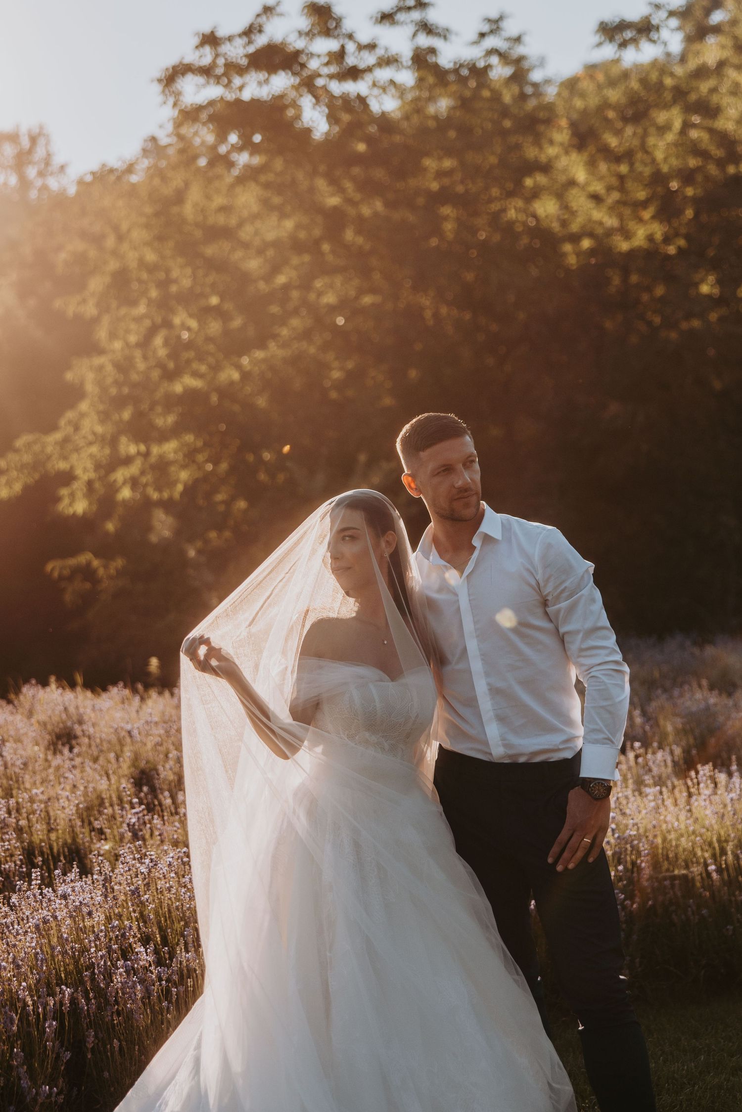 A newlywed couple posing in a lavender field at golden hour, bride in white gown with flowing veil.