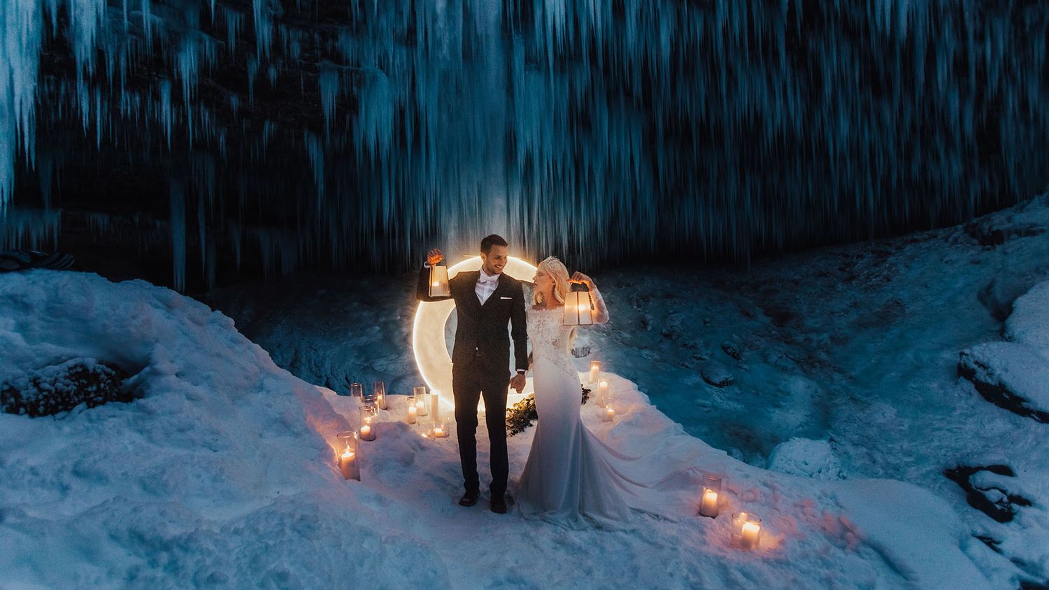 Dramatic winter wedding couple standing before a frozen waterfall with glowing candles and sparklers.