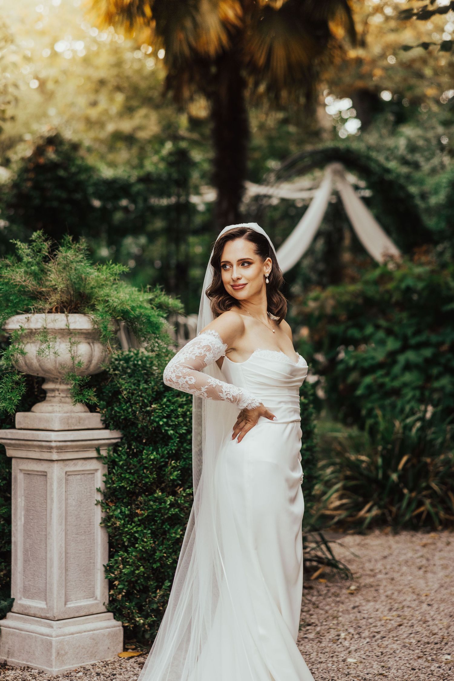 Elegant bride in white strapless gown with lace sleeves and veil posing in a lush garden setting.