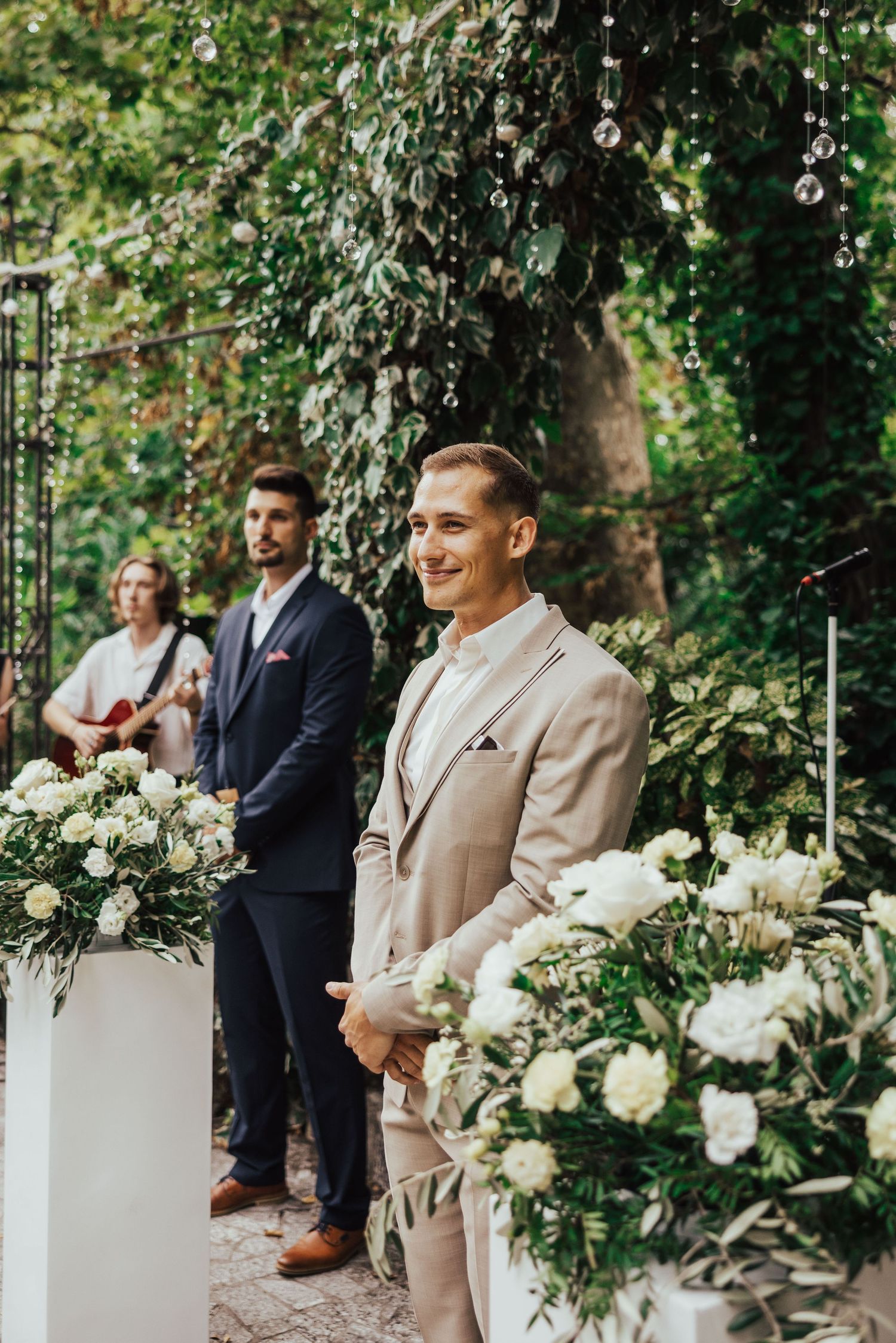 Groom in a beige suit smiles at the altar during an outdoor wedding ceremony with white floral arrangements and greenery.