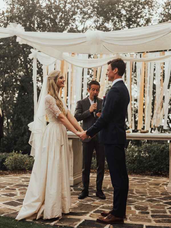 Bride and groom exchange vows under white draped outdoor wedding altar with officiant.