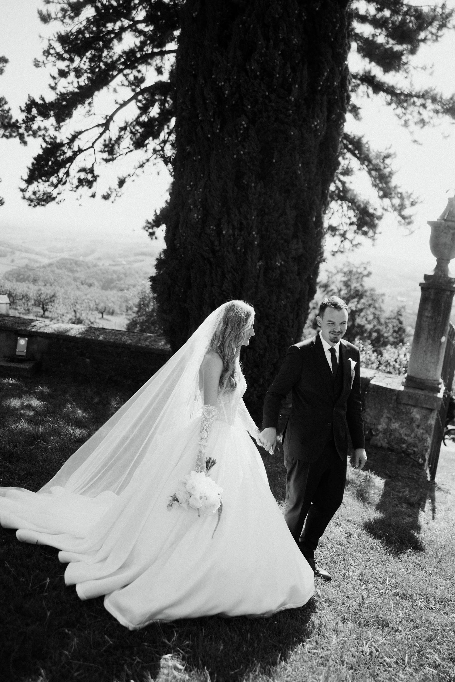 Black and white photo of bride and groom walking hand in hand near a large tree.