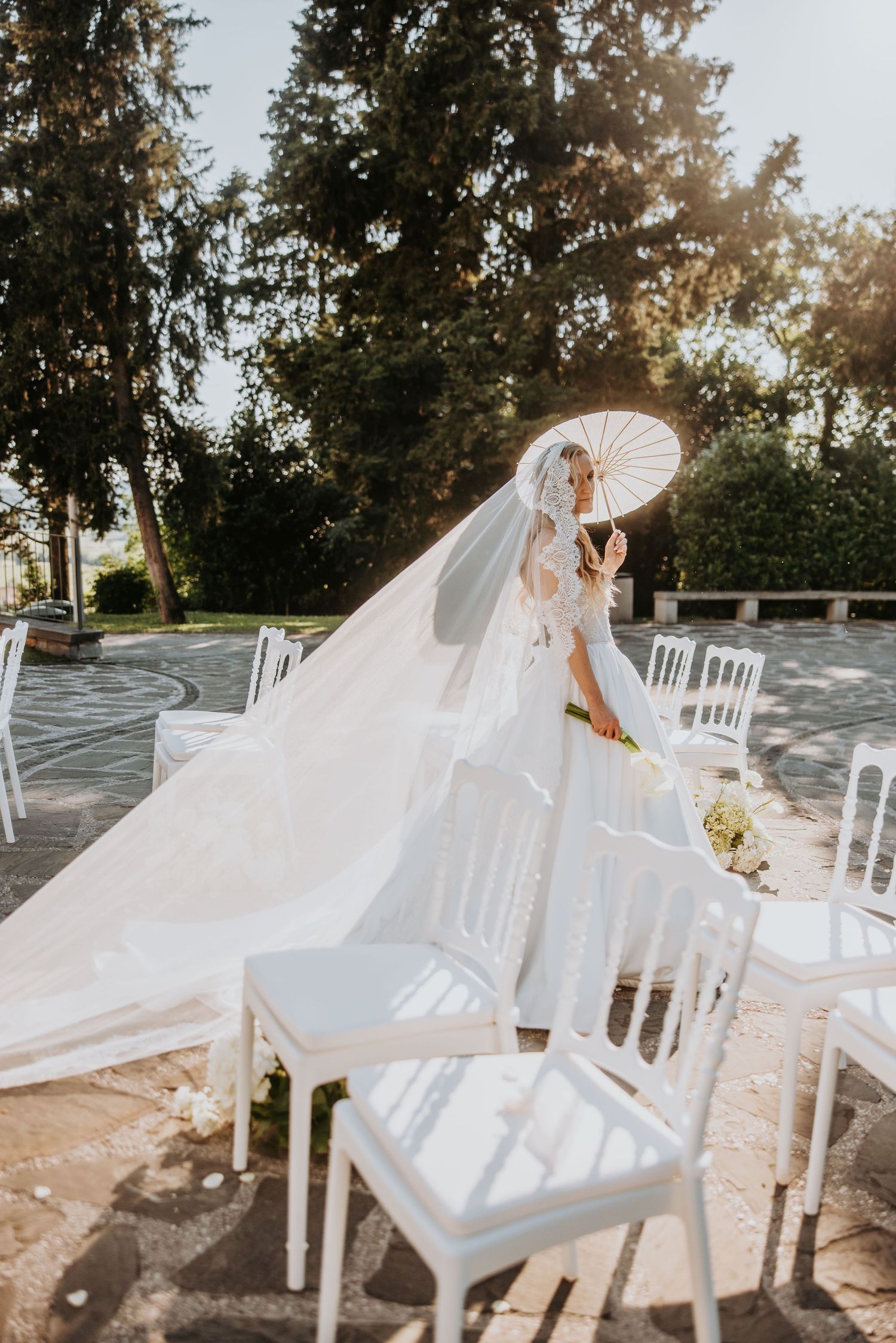 Bride in flowing white gown and veil holds parasol between white chairs at outdoor ceremony.