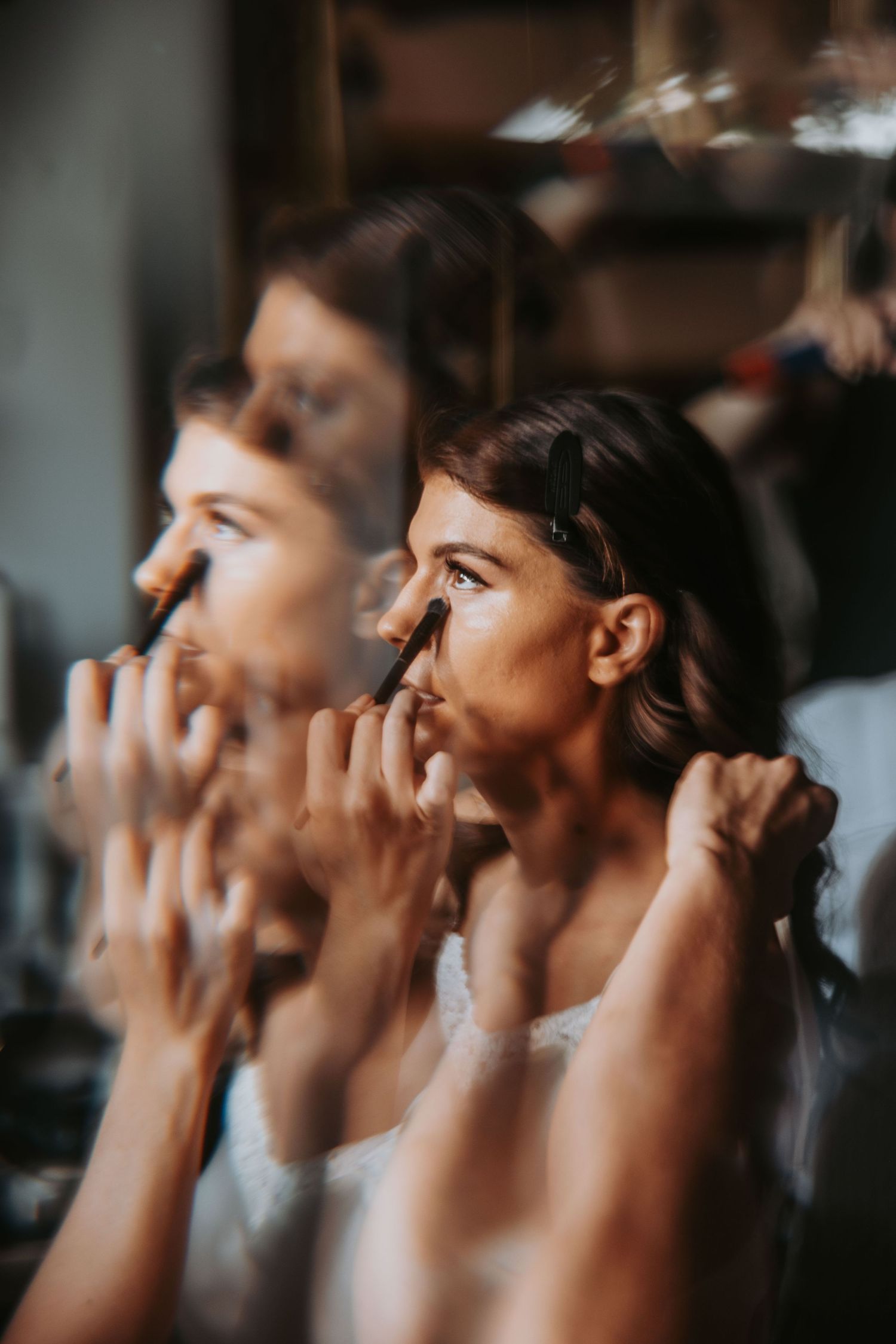 Bride applying makeup with a brush while gazing in mirror, elegant pre-wedding preparation moment.
