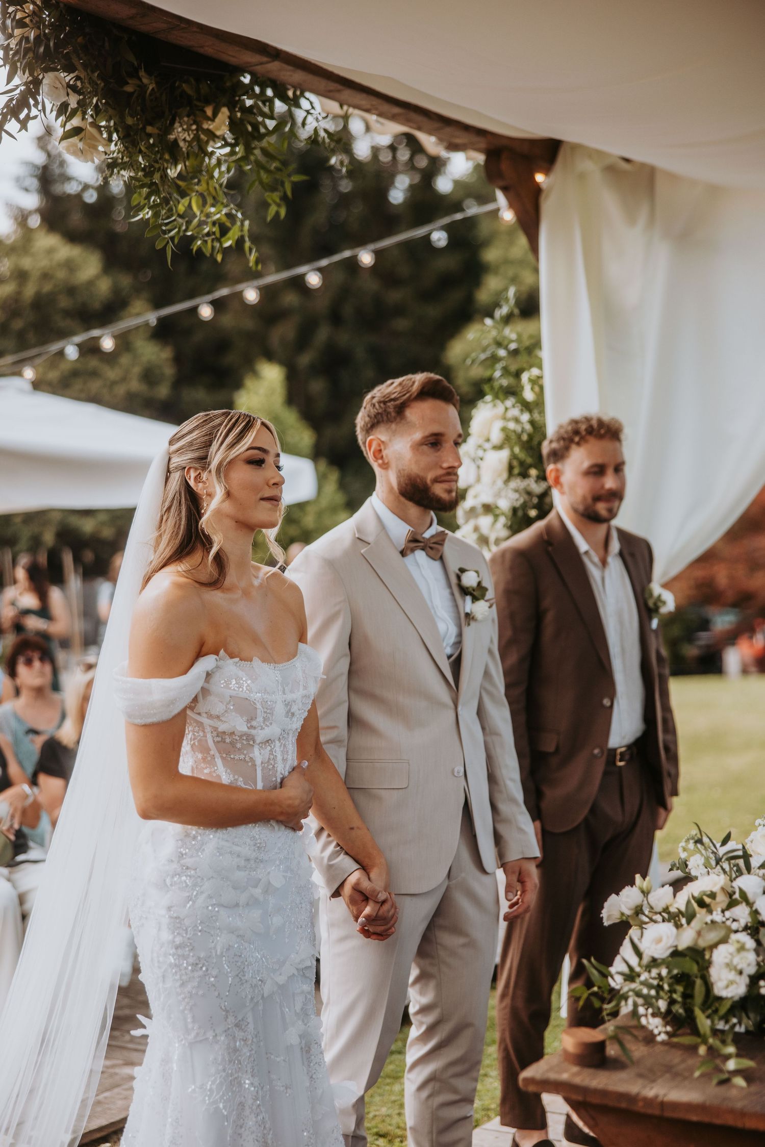 Bride and groom hold hands at outdoor wedding ceremony under floral arch with string lights.