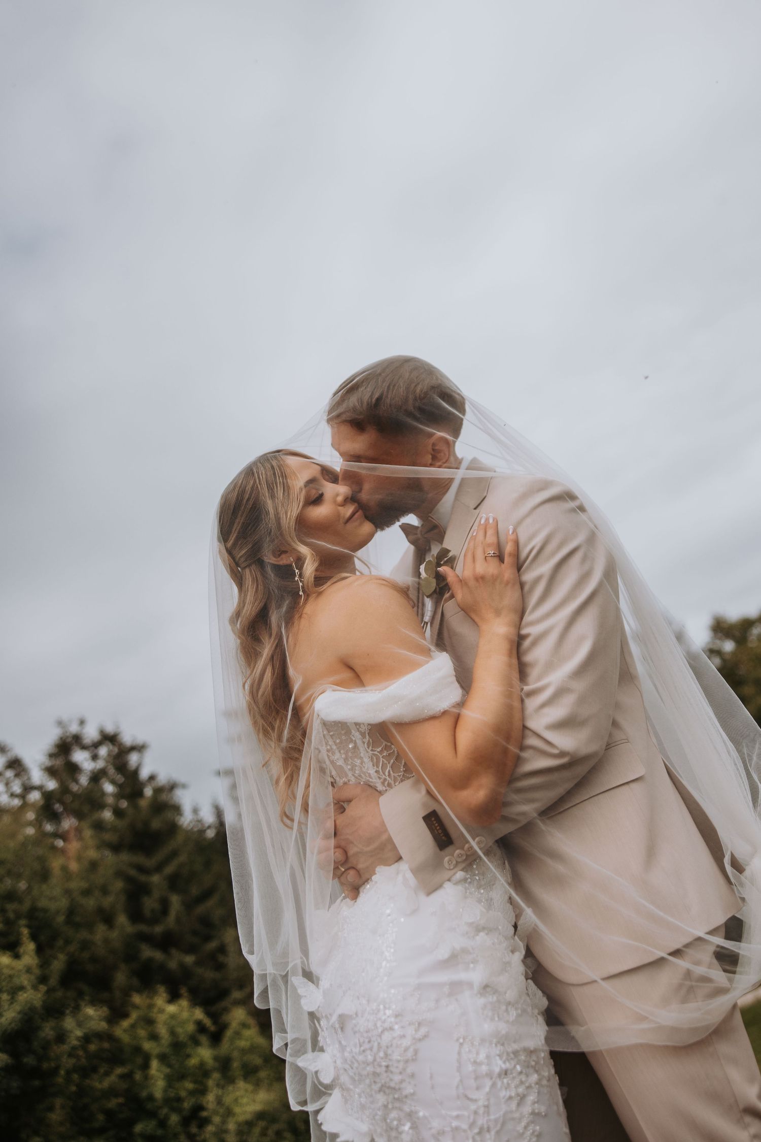 Bride and groom kissing under flowing veil against cloudy sky and green trees.