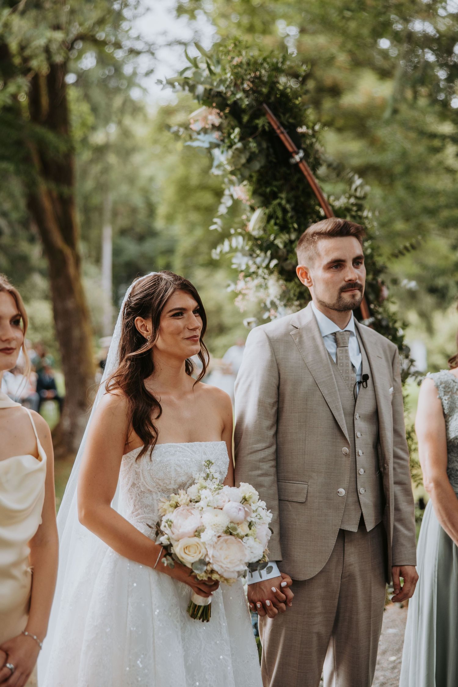 Bride holding a white bouquet stands beside her groom in a beige suit during an elegant outdoor wedding ceremony.
