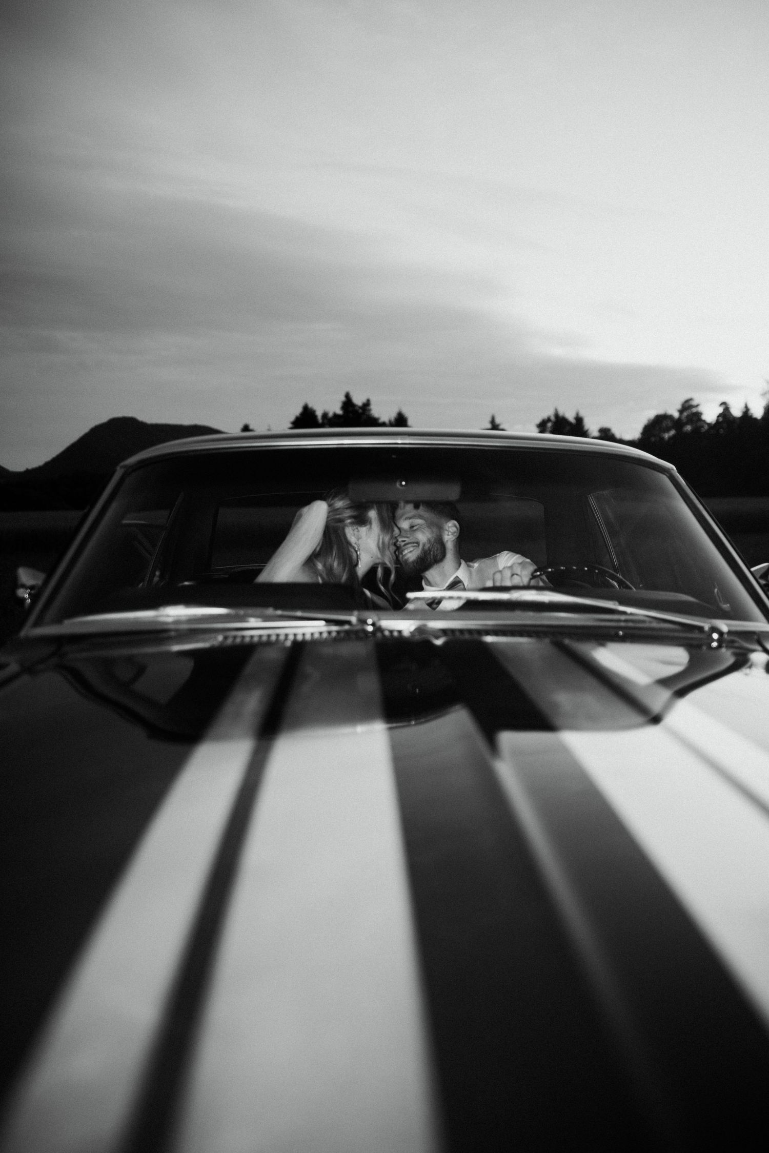 Black and white photo of newlyweds sharing a kiss inside a classic striped Mustang at dusk.
