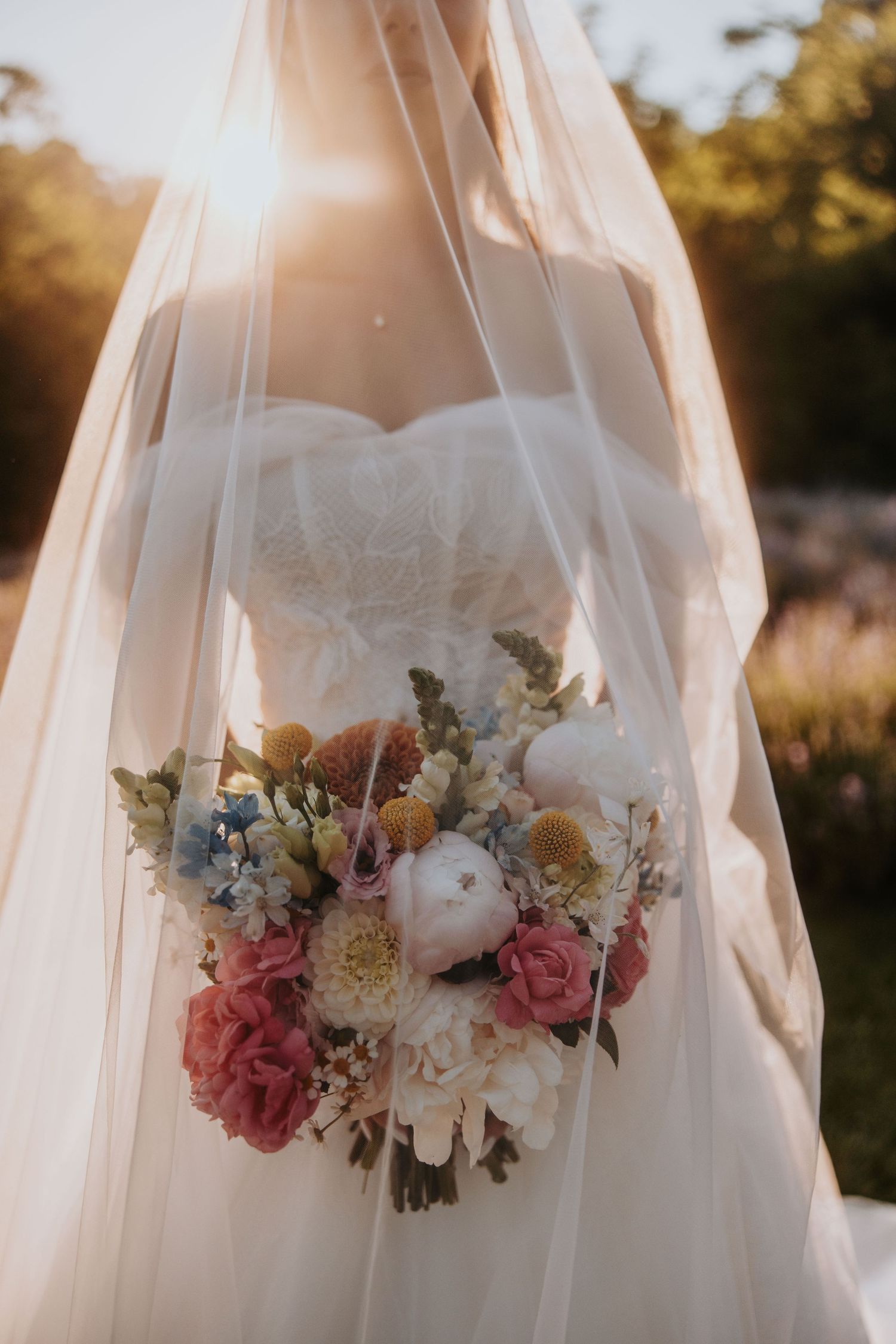 Close-up of bride holding colorful bouquet with peonies and wildflowers in golden sunlight.