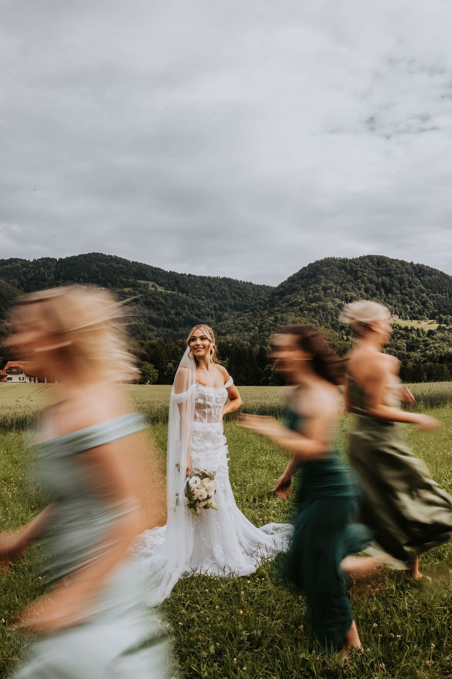 Bride in lace gown with veil standing in green meadow while bridesmaids run past in motion blur.