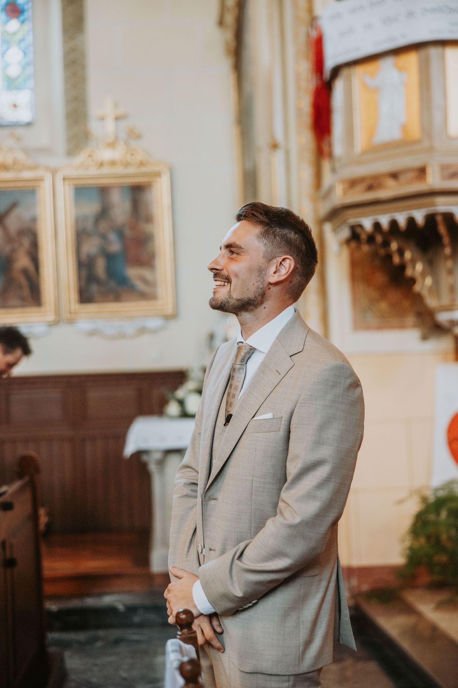 Smiling groom in beige suit standing at church altar awaiting bride during wedding ceremony.