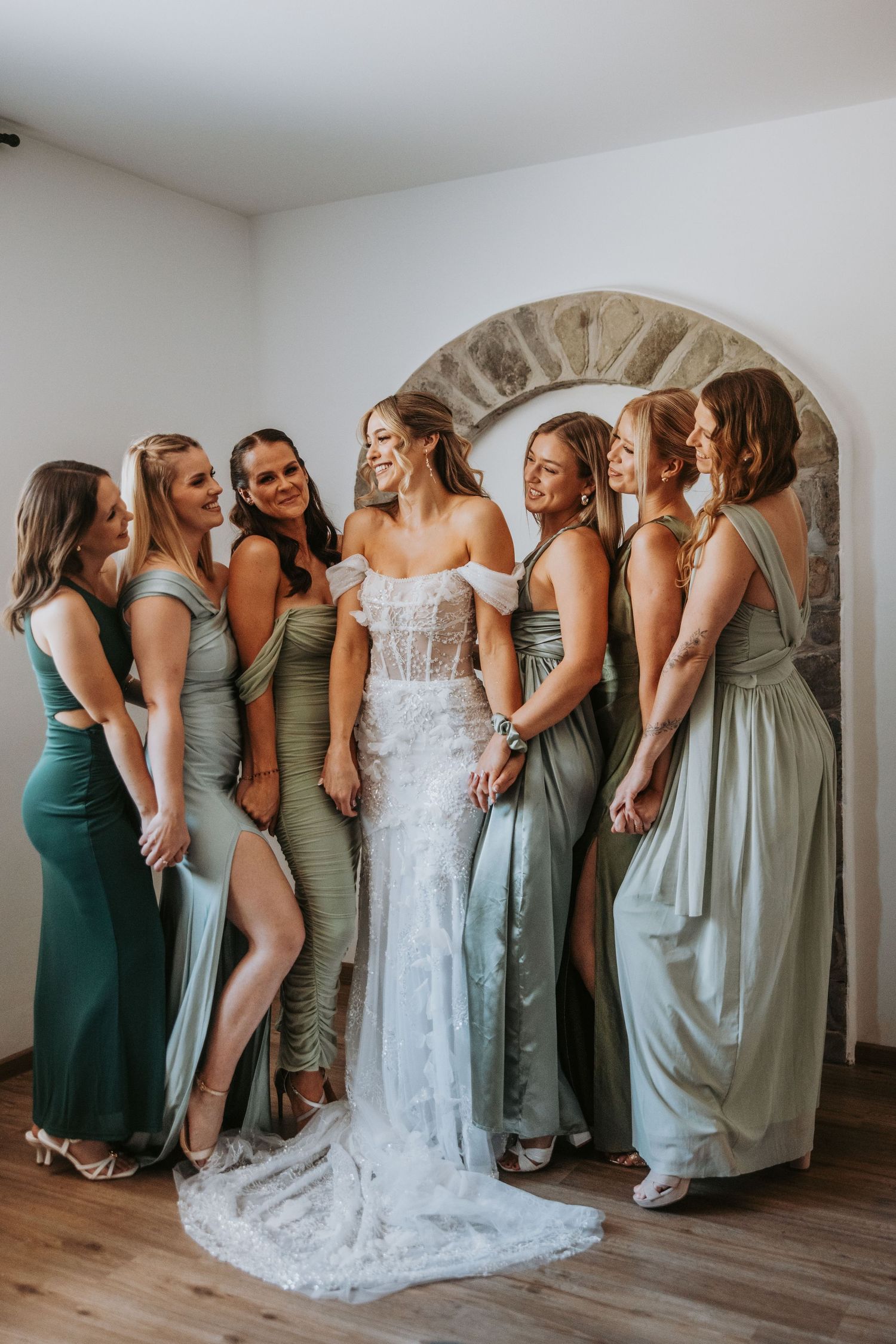 Bride in lace wedding gown laughing with six bridesmaids in sage green dresses near stone arch.