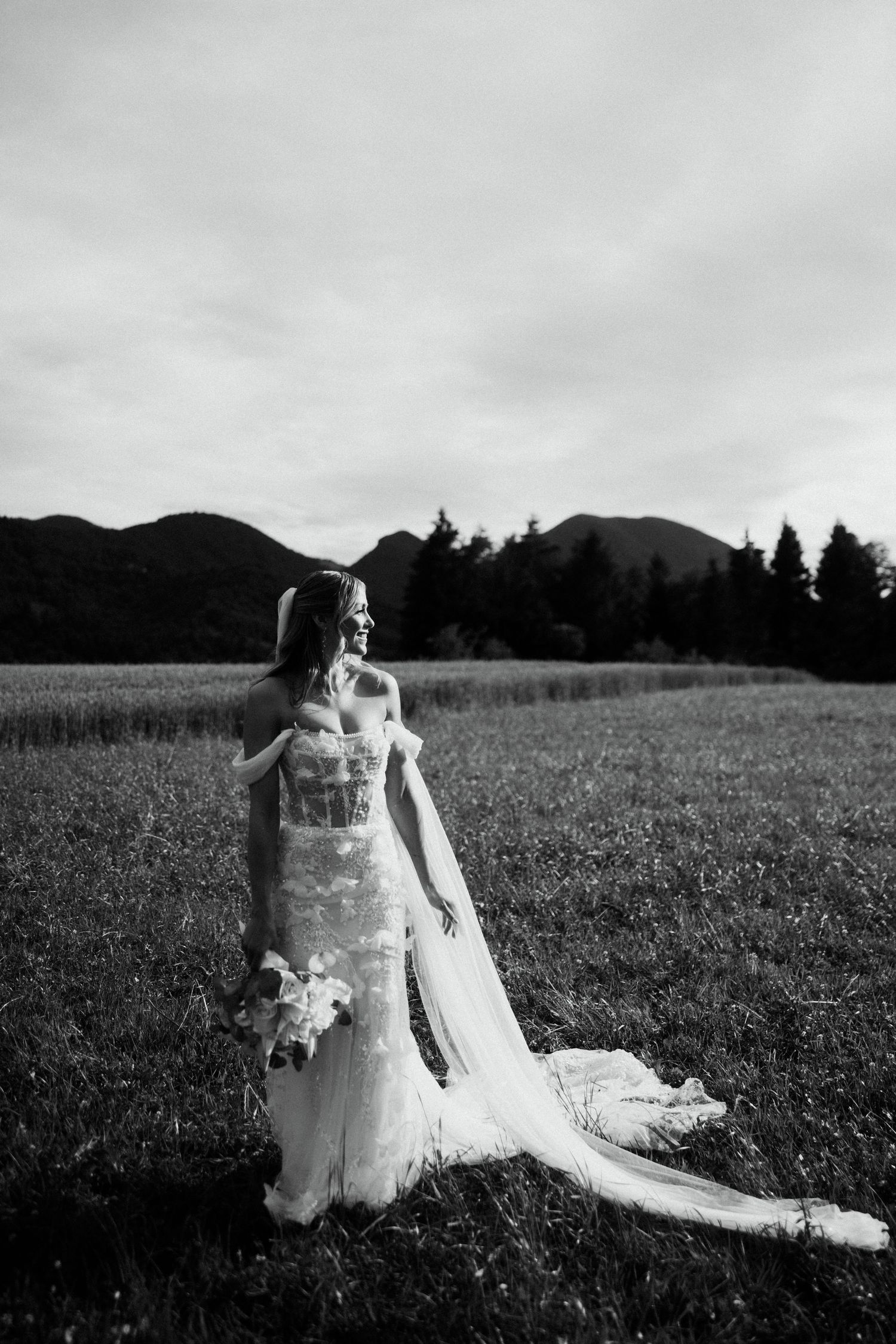 Black and white portrait of elegant bride in lace gown with flowing veil in scenic mountain meadow.