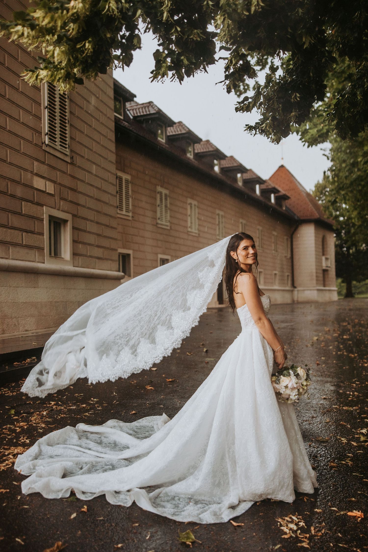 Bride in lace gown with billowing cathedral veil poses near historic stone building.