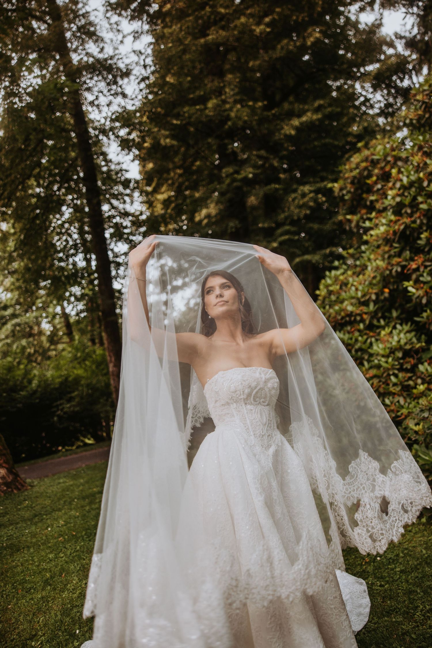 Bride in strapless lace gown lifts flowing veil gracefully in a lush garden setting.