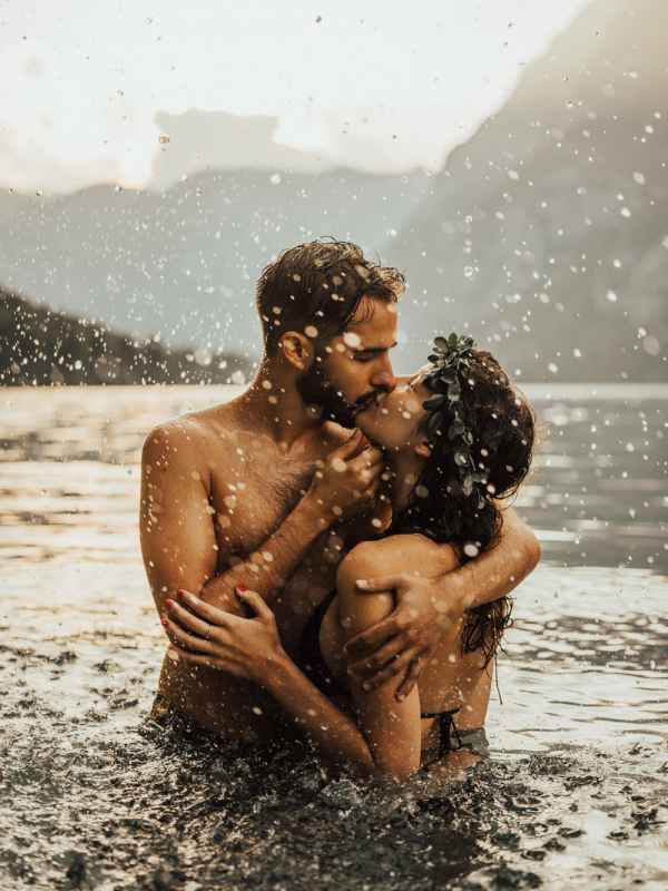 Passionate couple kissing in rain-splashed lake water with mountain backdrop in romantic photo.
