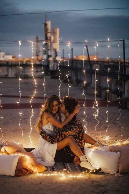 Romantic couple sitting on blankets surrounded by glowing fairy lights curtain at dusk in a cozy outdoor setting.